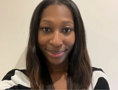 A person with long hair wearing a black and white striped top, standing against a plain light-colored wall.