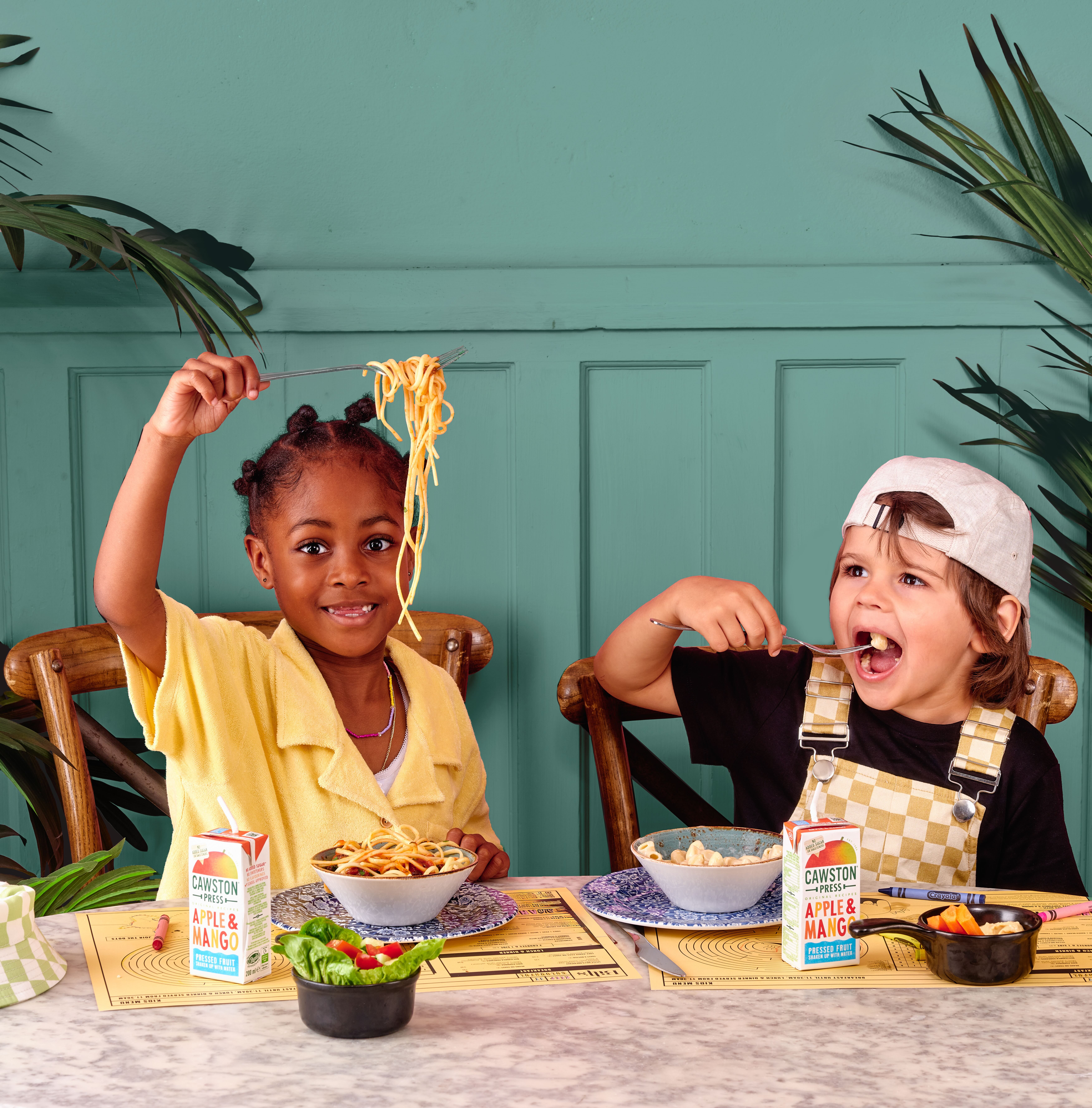 a boy and a girl sitting a table with a bright green wall behind them, the girl is holding up a fork with spaghetti and the boy is putting a fork with food into his mouth