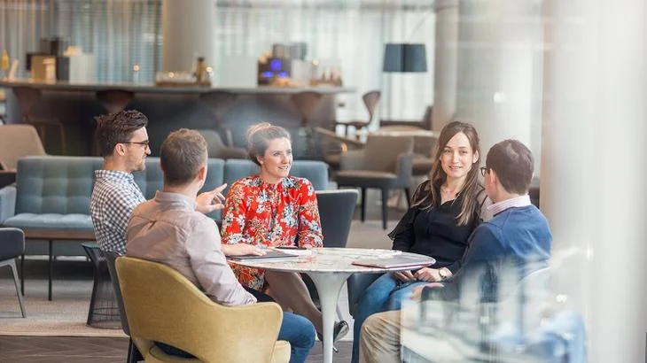 A group of five professionals collaborating at a round table in a modern office lounge area.