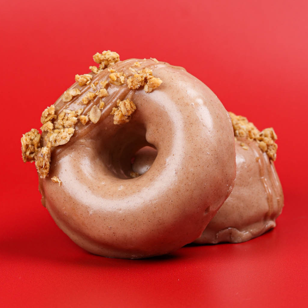 A close up of two doughnuts on a red background glazed and sprinkled with granola on one third 