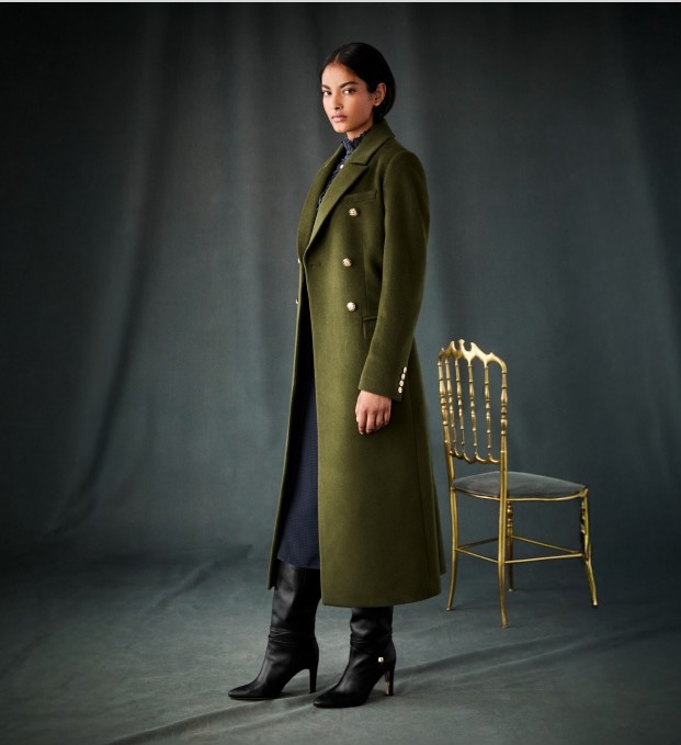 A woman wearing a long, green coat standing alone in an empty room with a chair