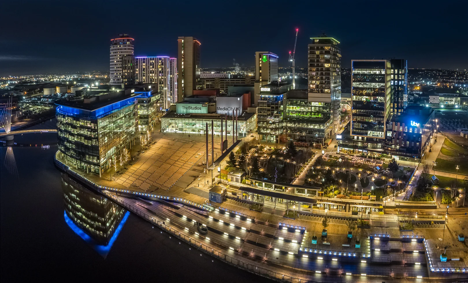Nighttime cityscape with illuminated modern buildings, a river, and a pedestrian bridge, creating a vibrant urban atmosphere.