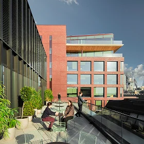 An **outdoor terrace** on a contemporary office building with red brick and dark metal facades, featuring potted plants and three people relaxing and talking.