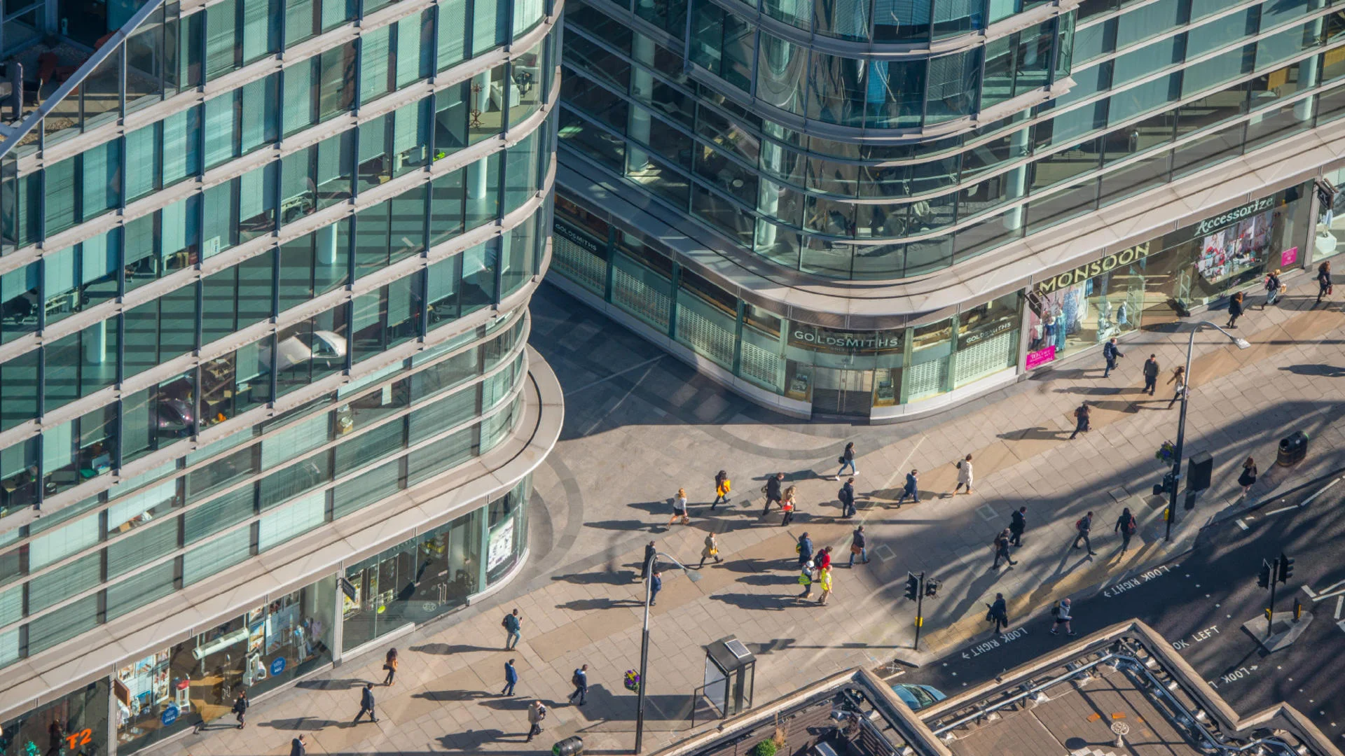 Aerial view looking down at a busy **London street-level retail area** surrounded by curved glass office buildings, with shops like **Goldsmiths** and pedestrians crossing the plaza.