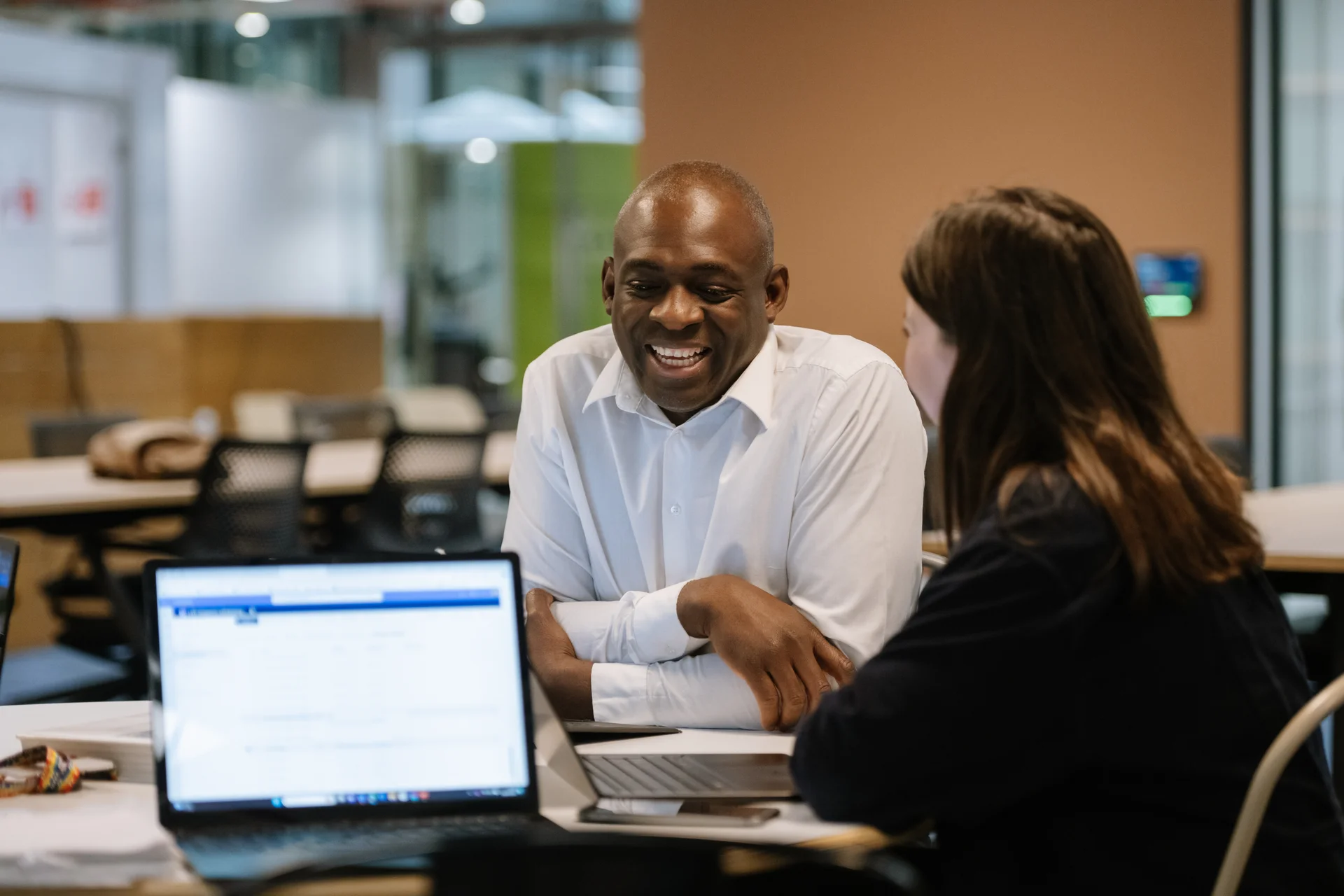Two individuals engaged in conversation at a desk with laptops, in a modern office setting with warm lighting.