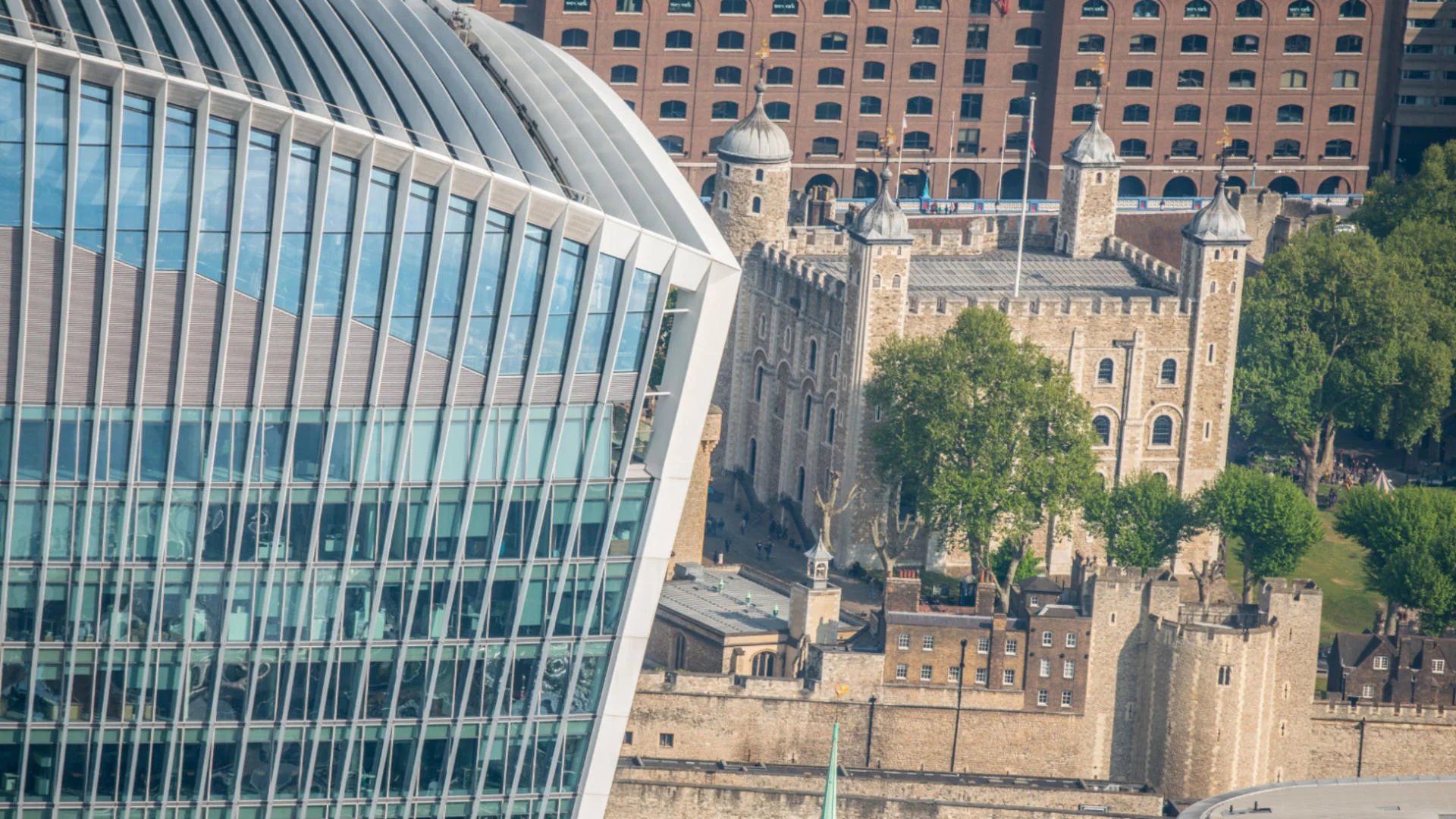 Modern glass building in front of historic stone castle with towers and greenery.