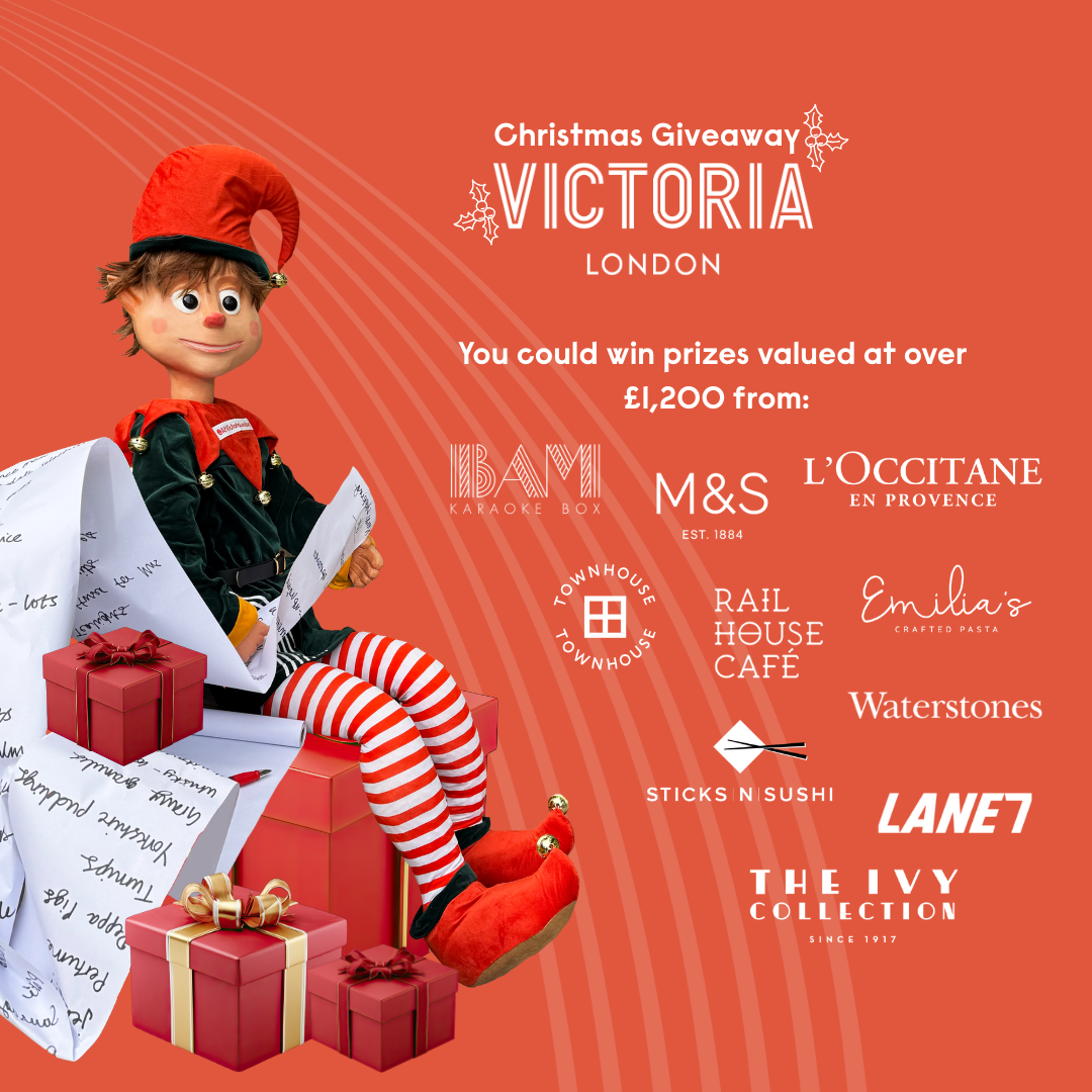 A Christmas elf sitting on a stack of presents holding a Christmas list next to white logos of At Victoria London retailers
