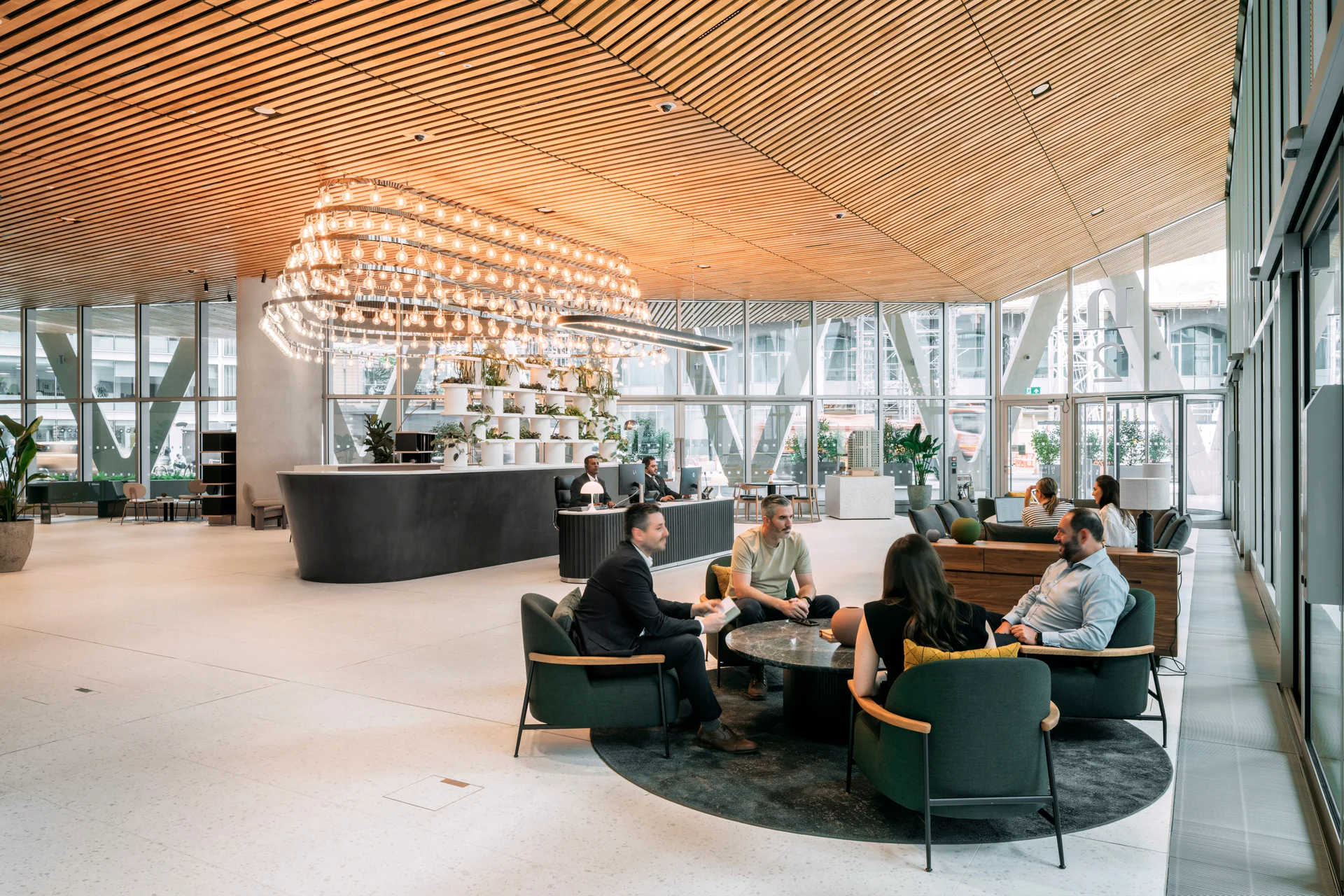 Modern office lobby with wooden ceiling, large windows, a chandelier, and people seated in a casual meeting area.