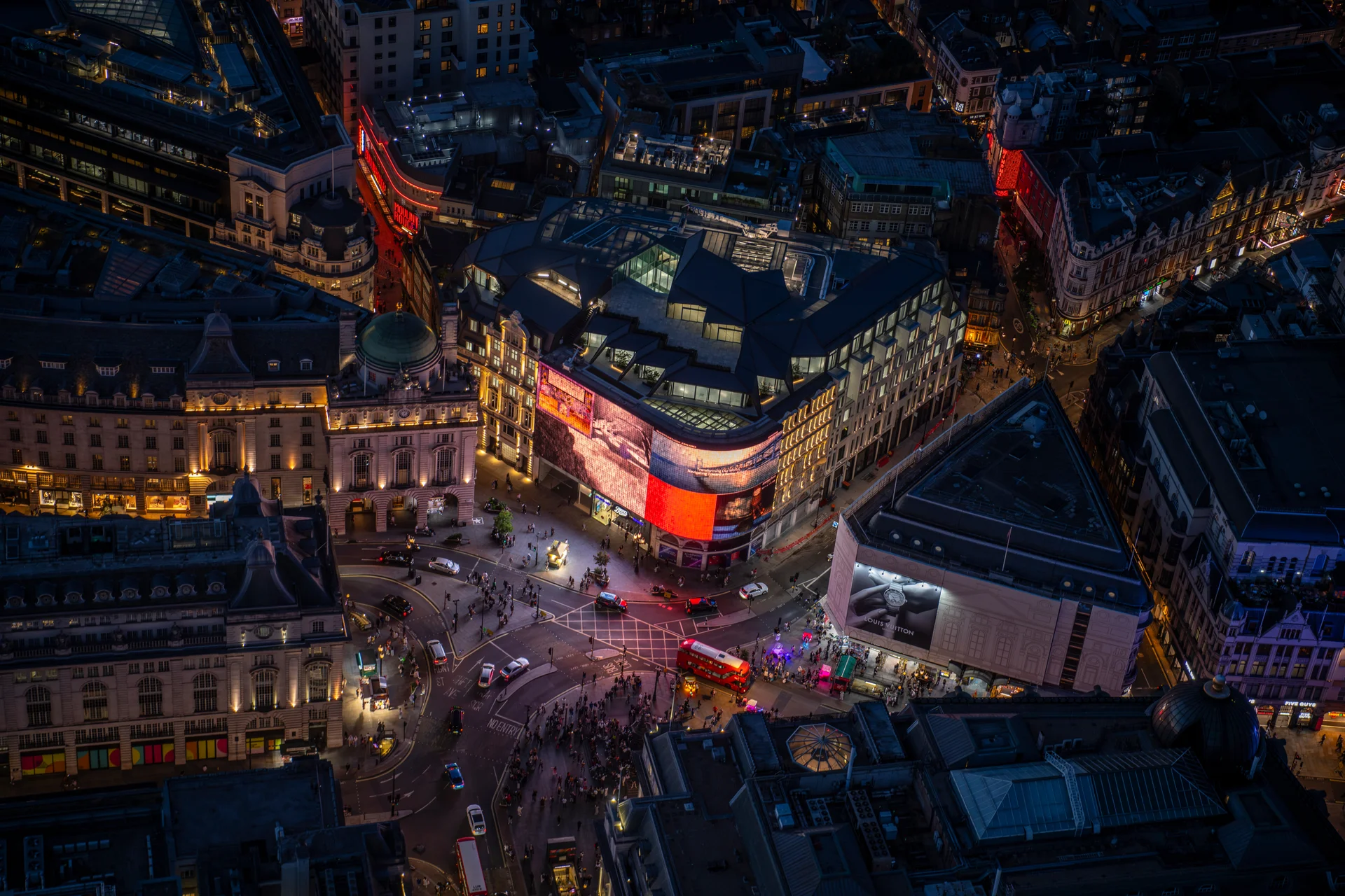 Aerial night view of a bustling urban intersection with illuminated buildings, pedestrians, and vehicles.