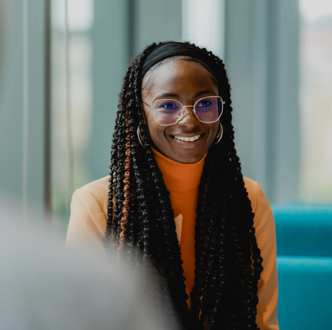 Smiling professional Black woman with long braids and round glasses, wearing an orange turtleneck in a modern office or meeting setting.