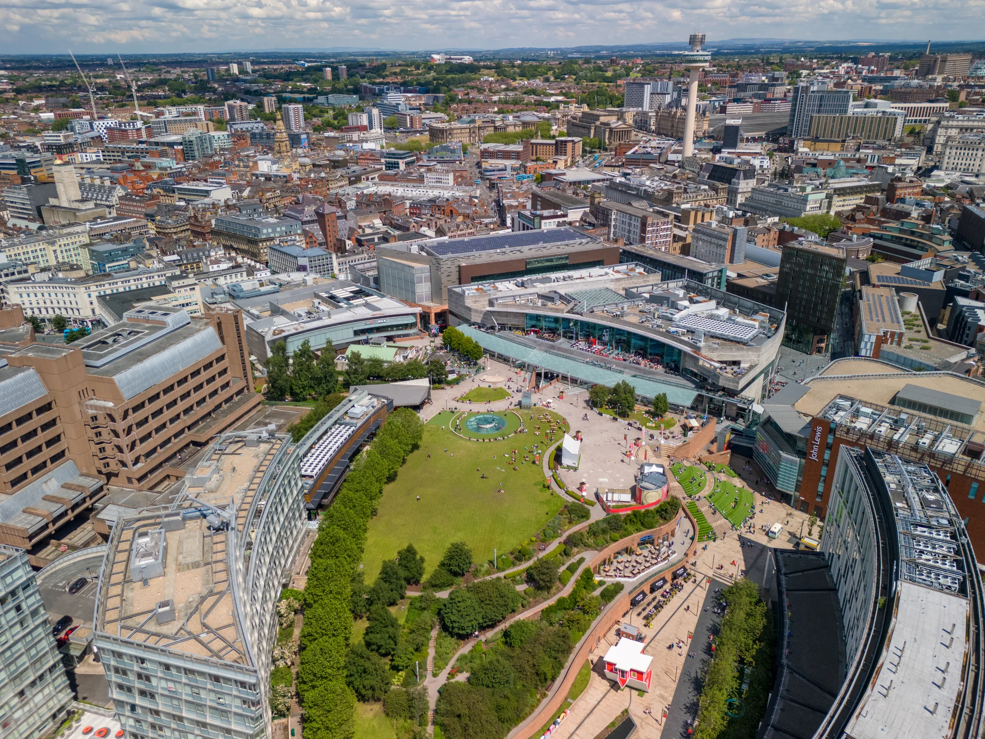 Aerial view of a cityscape with modern buildings, a central green park, and people enjoying outdoor spaces on a sunny day.