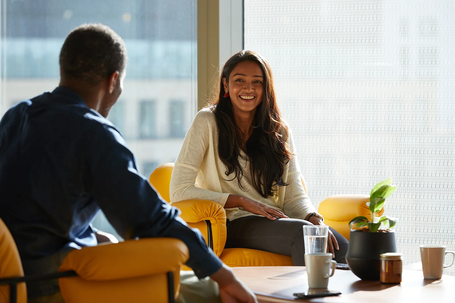 Two people engage in conversation in a modern office setting with yellow chairs, a table, and natural light from large windows.