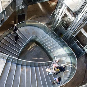 Overhead view of a dramatic, **curved geometric staircase** with glass railings inside a modern commercial building atrium, with people walking and standing on the steps.