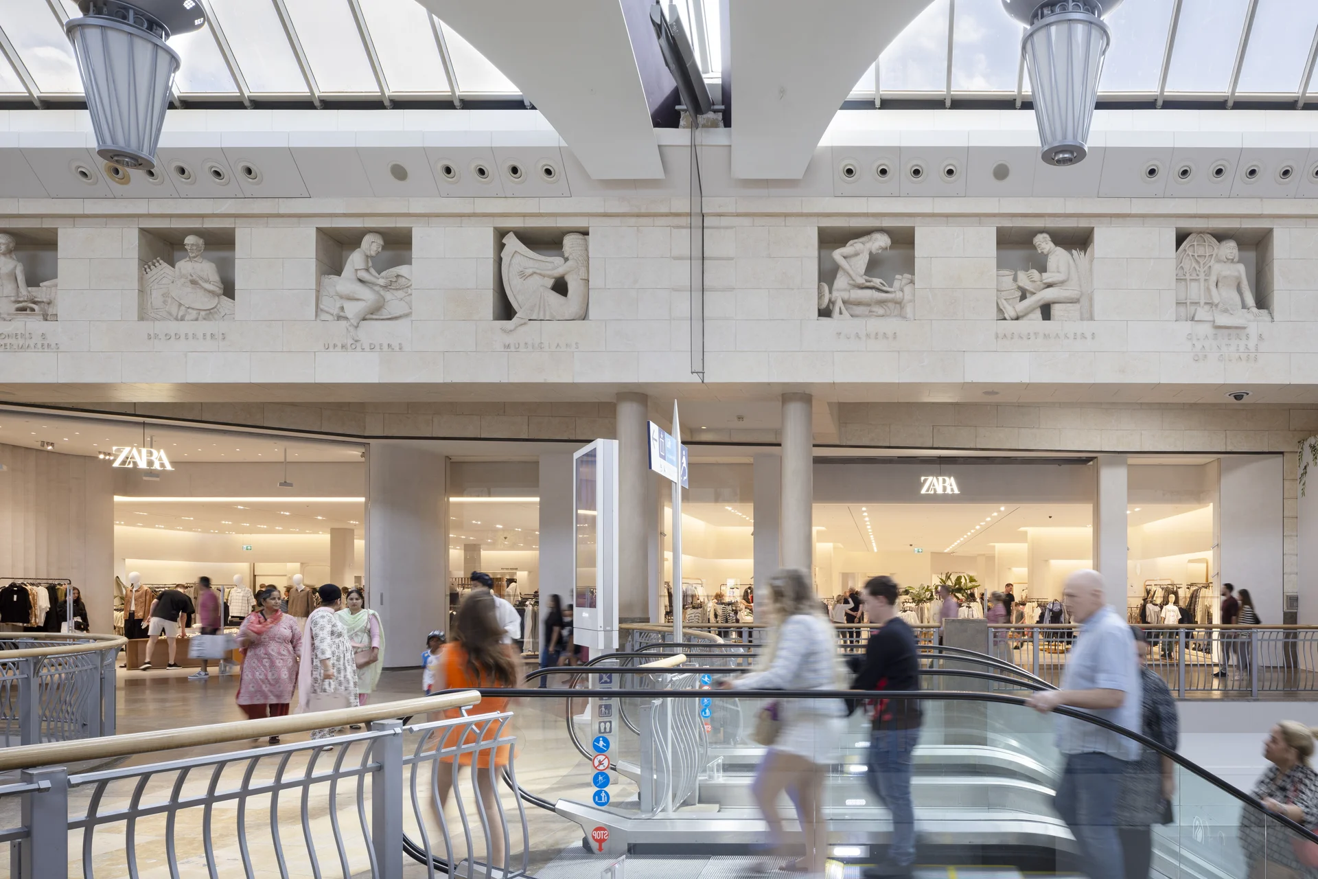 Interior view of the **Bluewater Shopping Centre** mall, highlighting the decorative stone relief sculptures depicting different trades above the storefronts, with shoppers on the walkway near the **Zara** store.