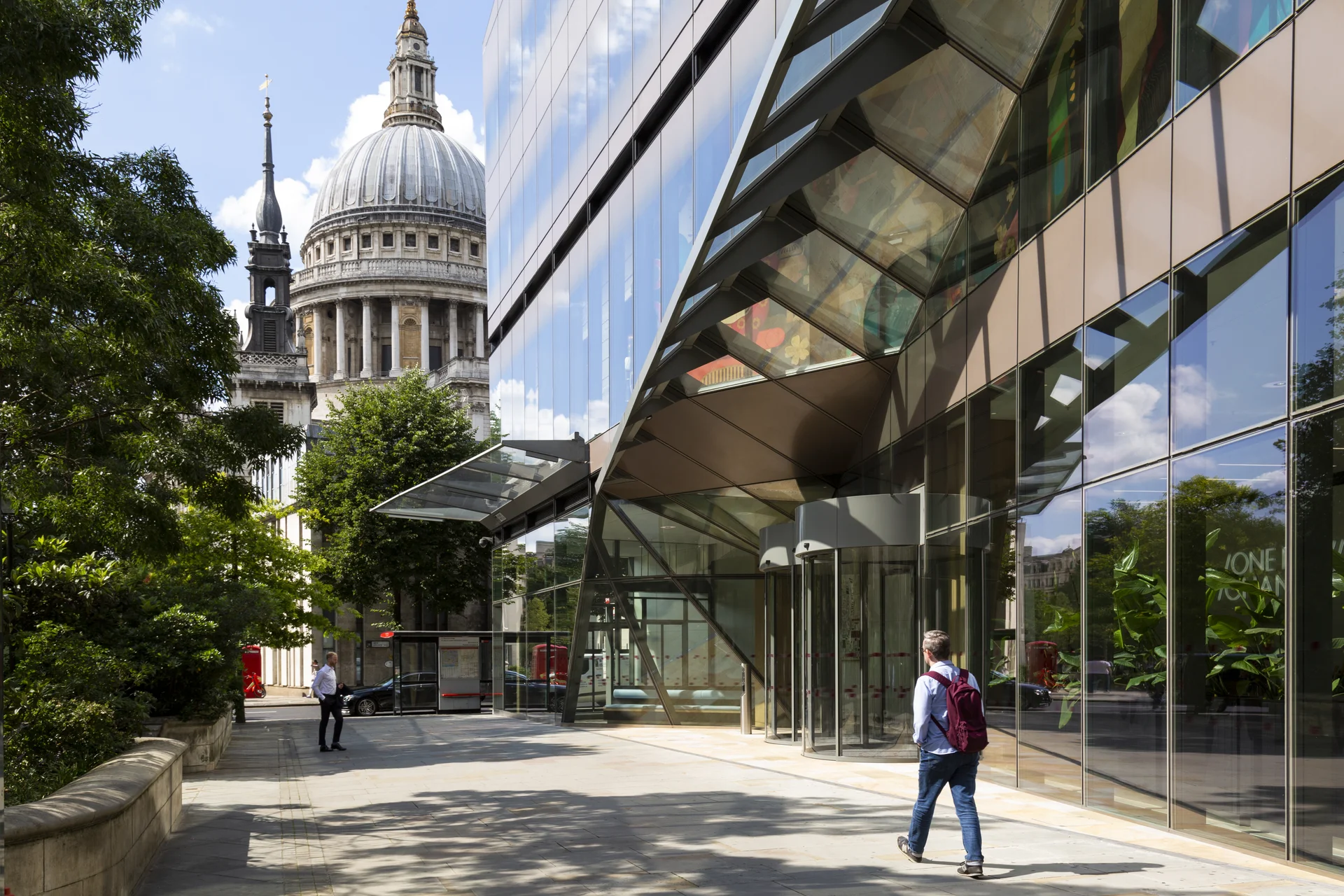 Architectural contrast in London: A modern glass office facade next to the historic dome and spire of **St Paul's Cathedral**, featuring a pedestrian walkway.