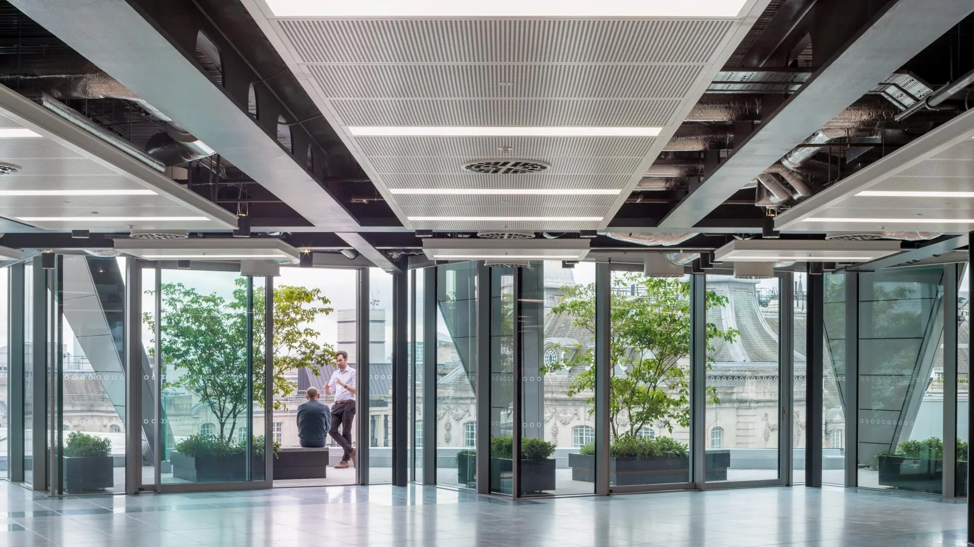 Modern interior with exposed beams, glass walls, and two people conversing, overlooking trees and a classical building.