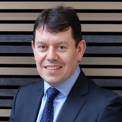 Close-up photo of a man with dark hair, wearing glasses, smiling for a corporate headshot with a striped wooden panel backdrop.