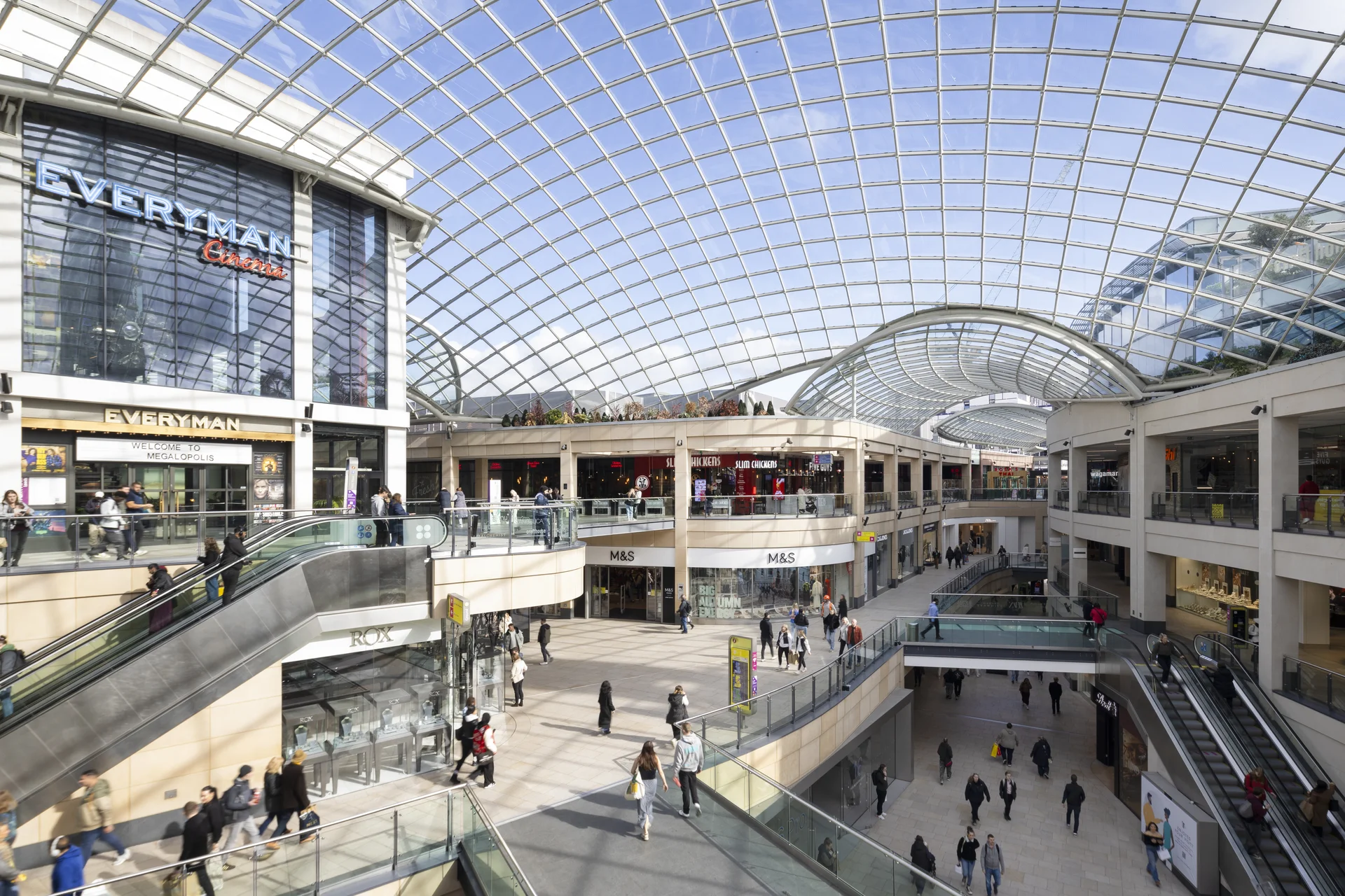 A bustling indoor shopping mall with a glass roof, multiple levels, escalators, and shoppers walking around.