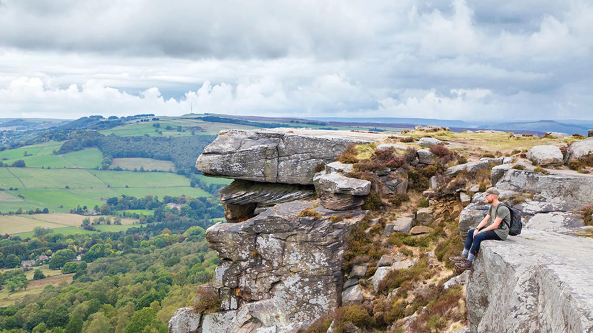 Person sitting on rocky outcrop overlooking green fields and forests under a partly cloudy sky.