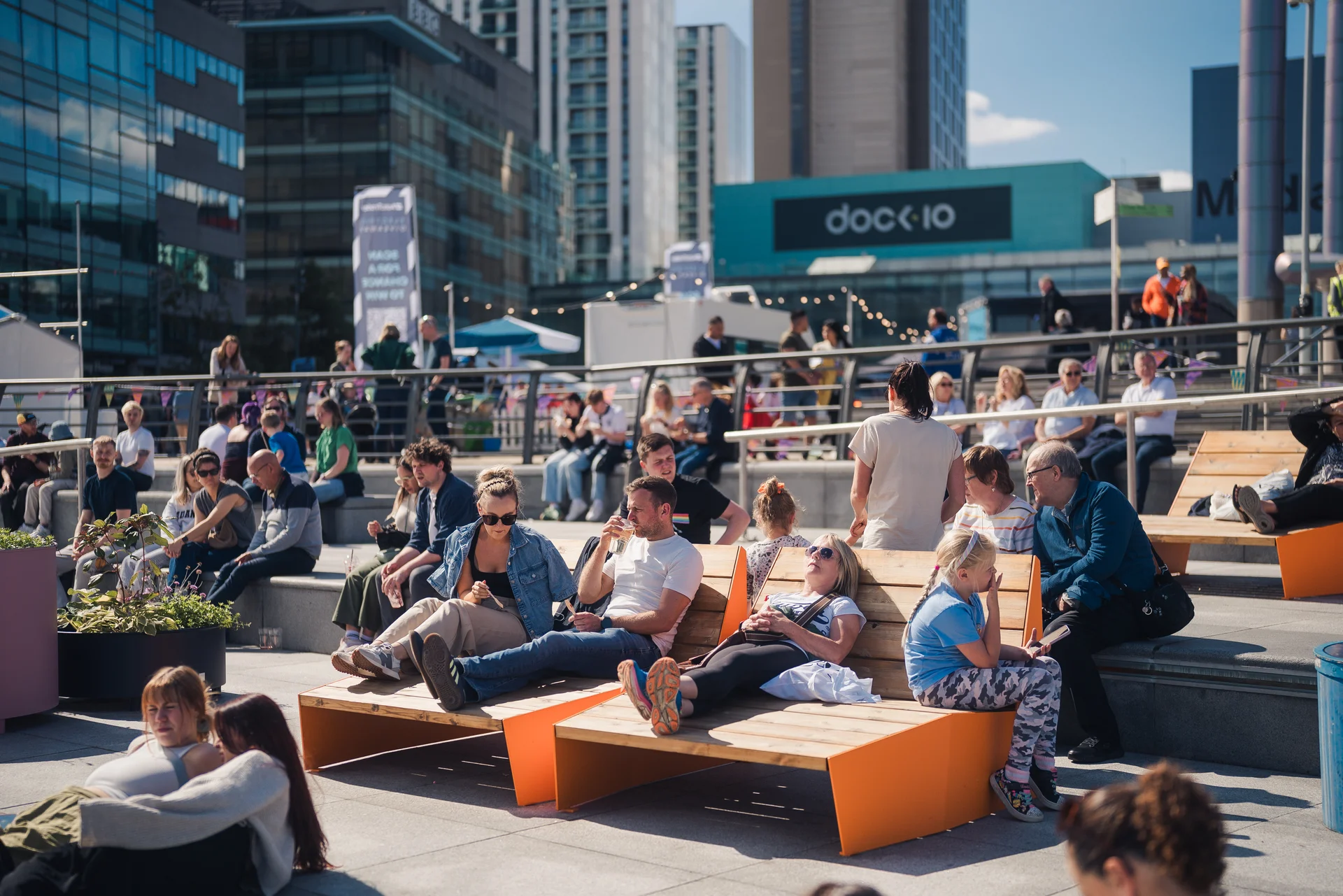 People sitting on orange benches on a sunny day at MediaCityUK, with the Dock10 sign visible on a modern glass building.