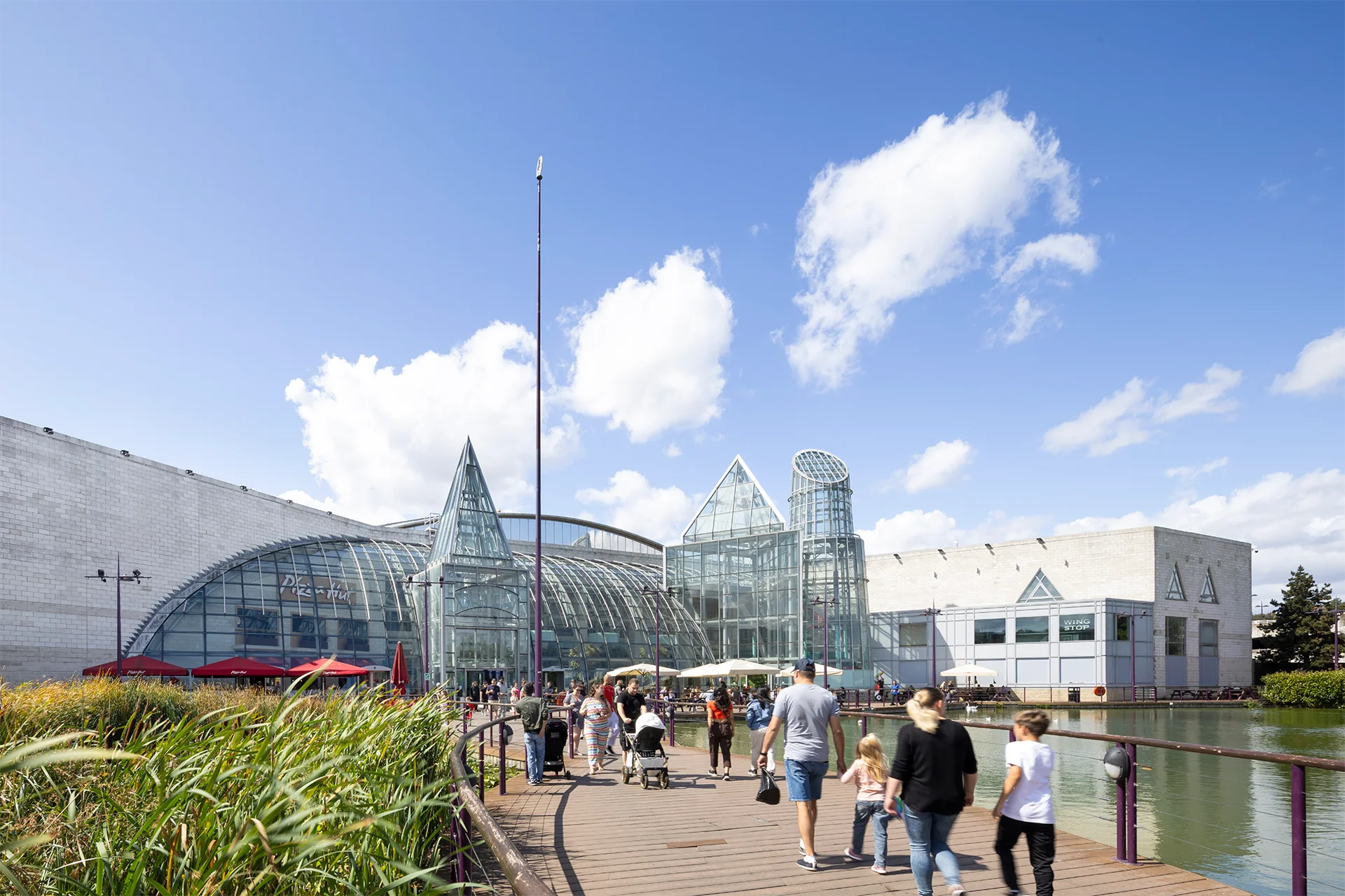 A sunny day at a modern complex with glass structures, people walking along a wooden path by a pond, and lush greenery nearby.