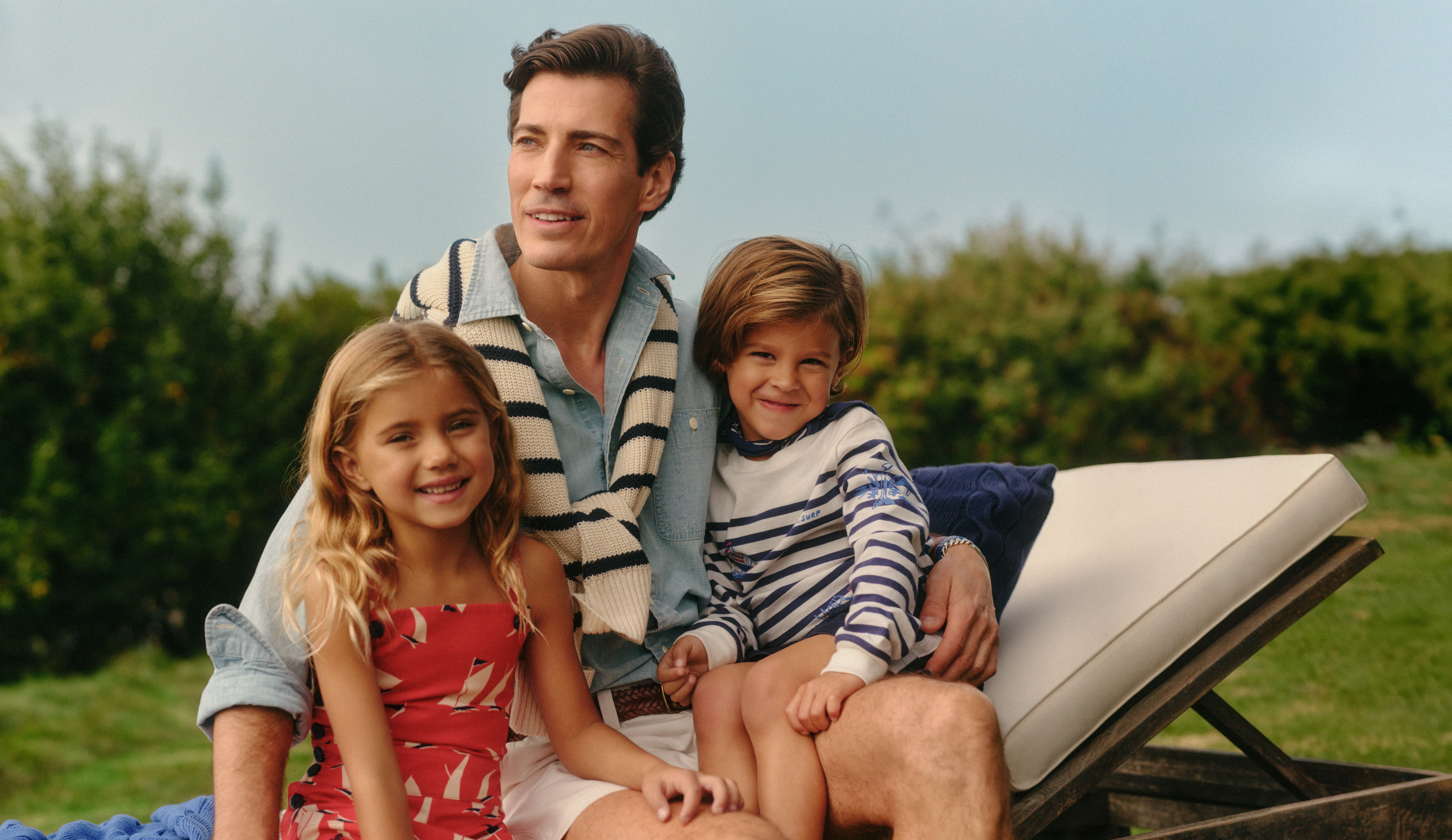 a man in a pale blue shirt sitting on a white sun lounger with a girl and a boy. The main has a white and navy stripe jumper tied around his shoulders. The girl is wearing a red dress and the boy is wearing a white jumper with blue stripes. Grass and a hedge can be seen in the background.