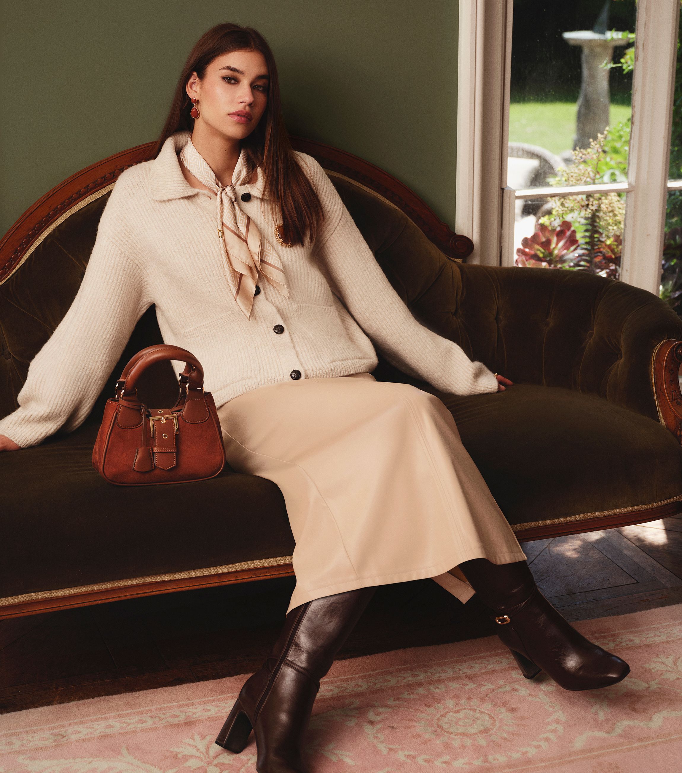A woman in beige clothes and brown boots lounges on a sofa.
