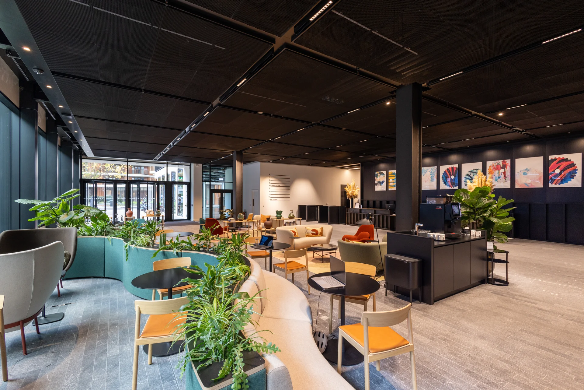 Modern lounge with curved seating, plants, tables, and colorful chairs under a dark ceiling with recessed lighting.