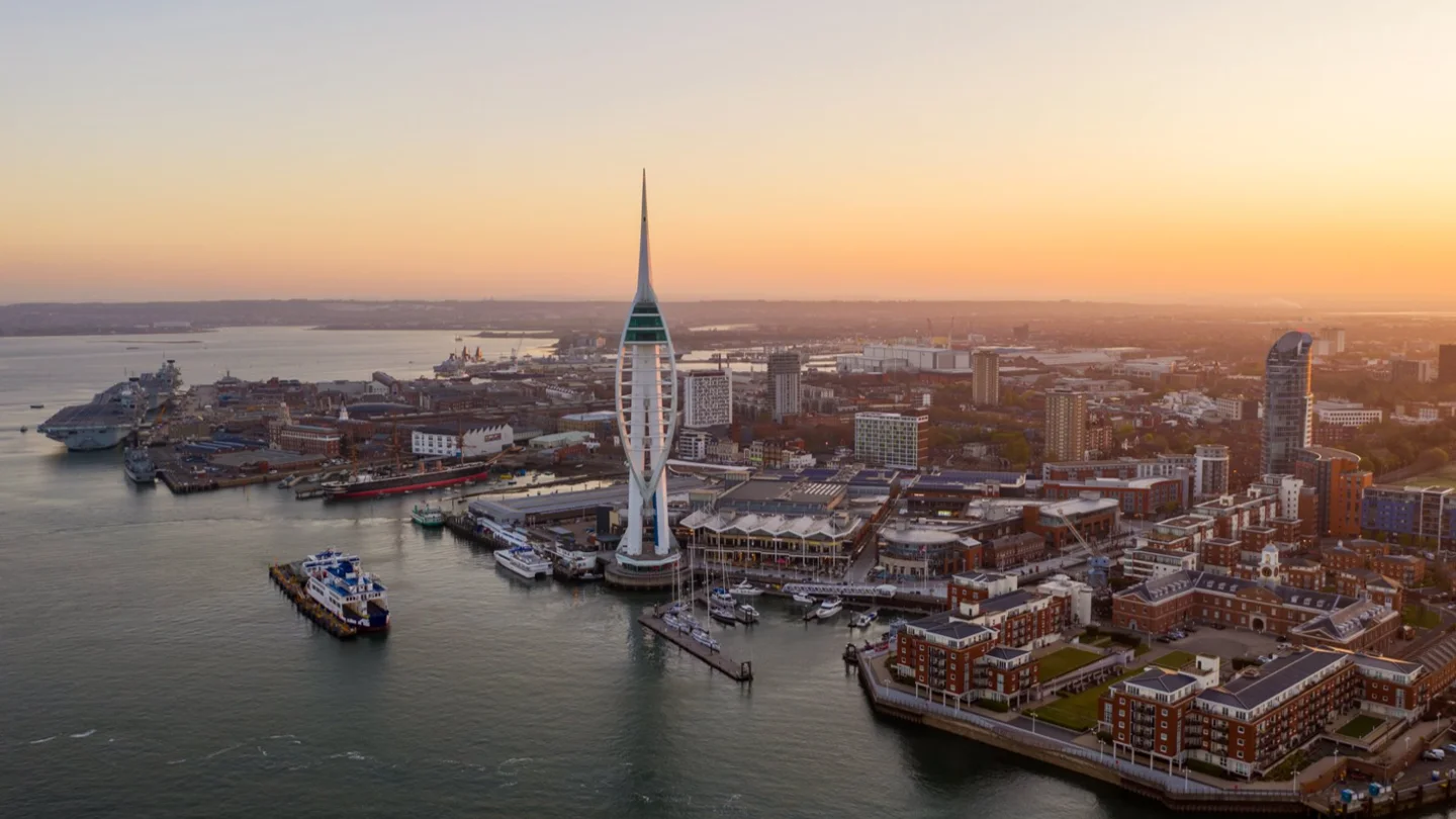 **Aerial view** of the **Portsmouth** cityscape and harbour at sunset, featuring the prominent **Spinnaker Tower** and the surrounding **Gunwharf Quays** retail and leisure complex.