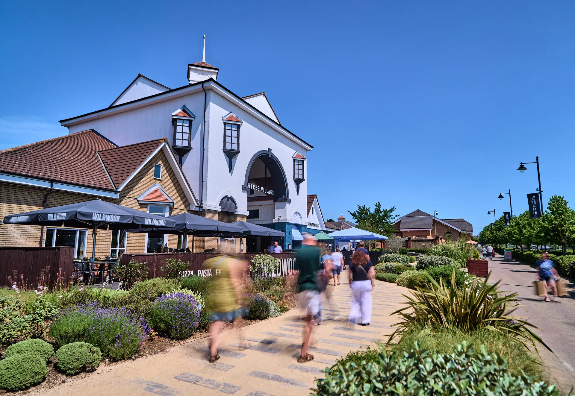 People stroll along a sunny path near a white building with a pitched roof, outdoor seating, and lush greenery.