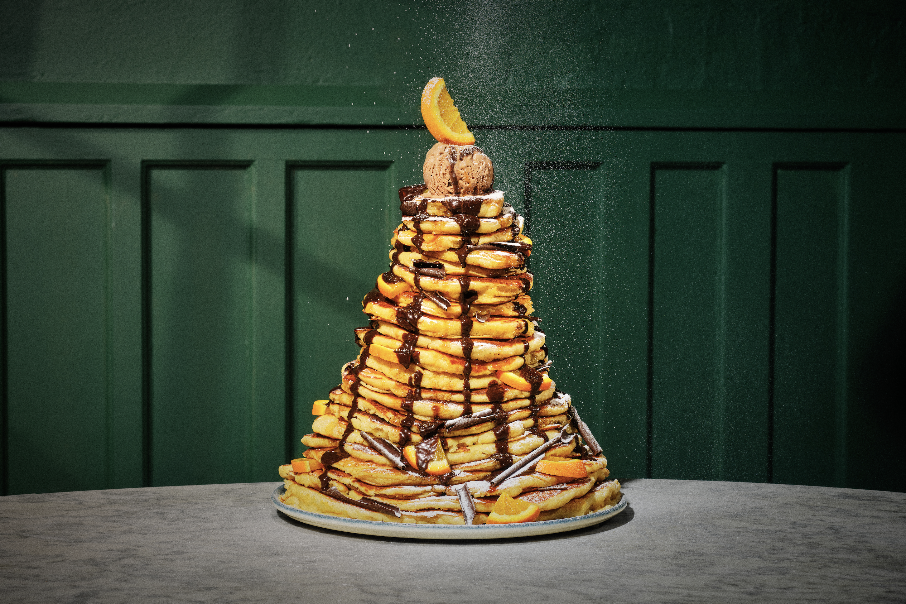 A large pyramid stack of pancakes topped with a scoop of ice-cream and garnished with a segment of orange
