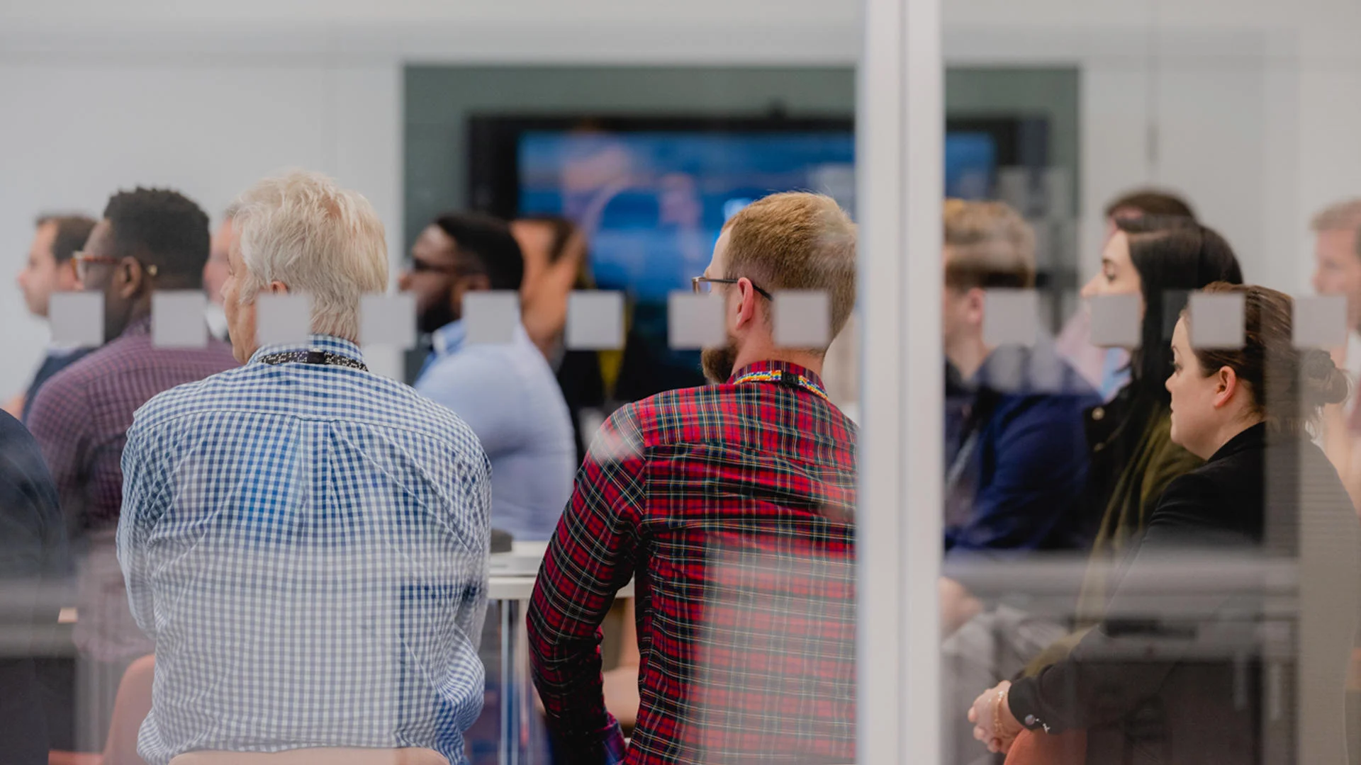 Group of people seated in a modern meeting room, seen through a glass wall.