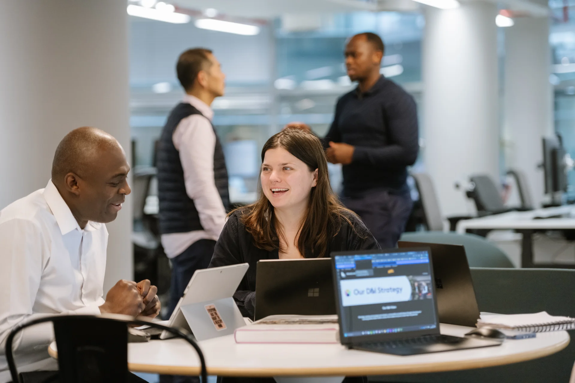Two people collaborate at a desk with laptops in a modern office, while two others stand in the background.