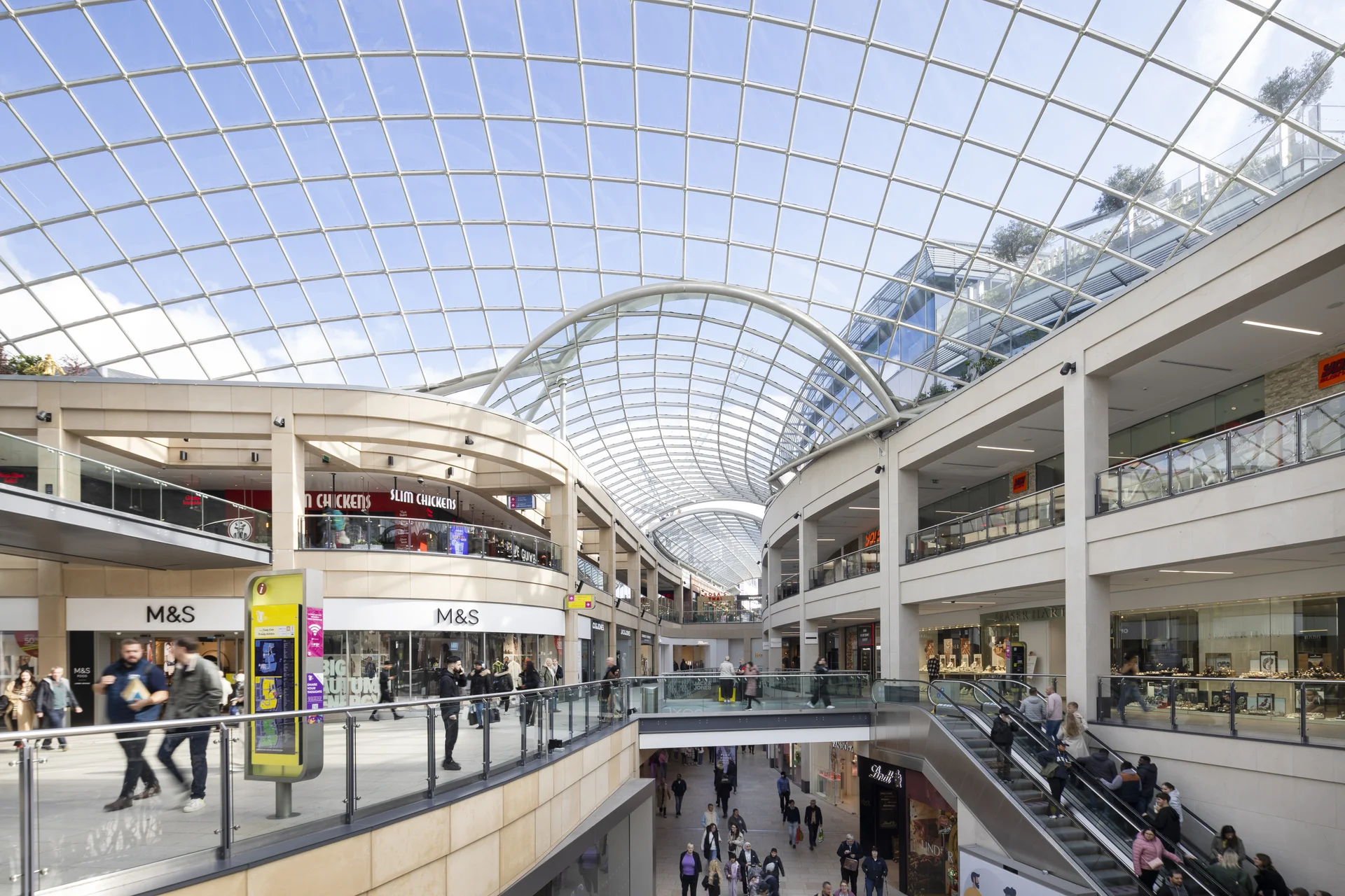 Wide interior of Trinity Leeds shopping centre. Features a large arched glass roof and multiple levels of stores like M&S.