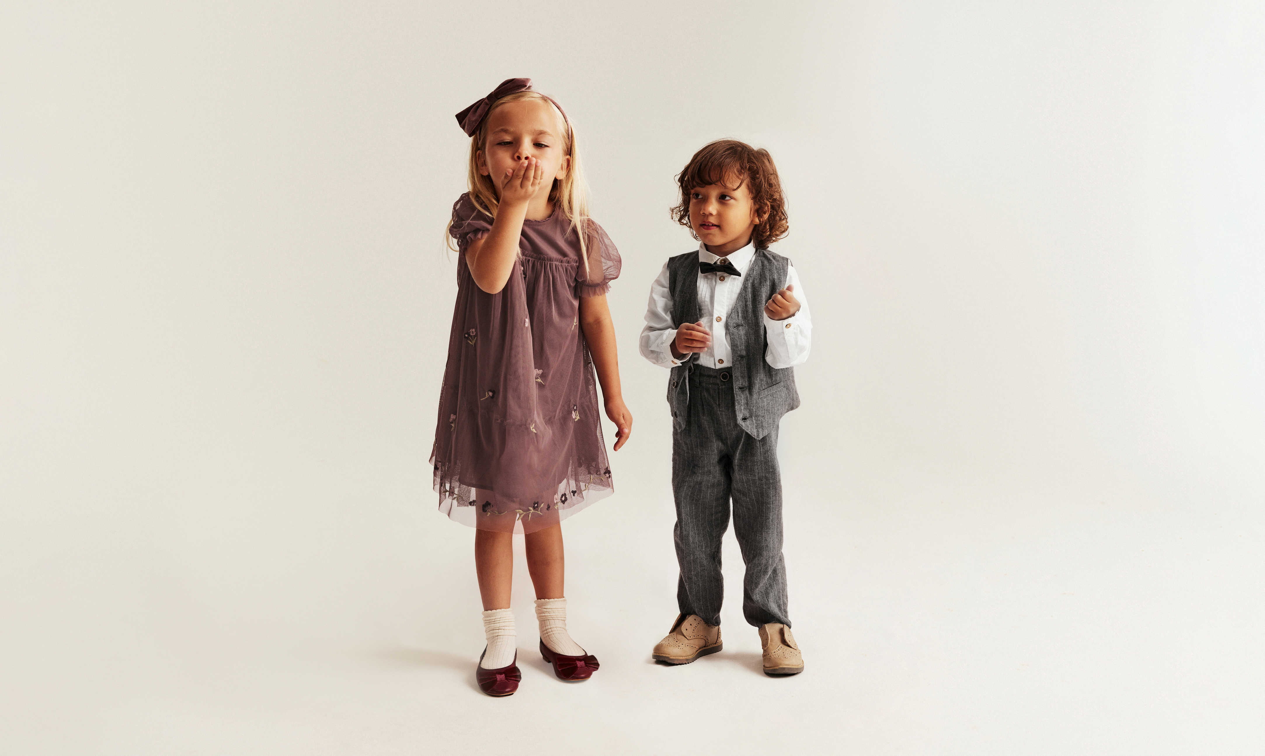 Two children in partywear stood against a beige background