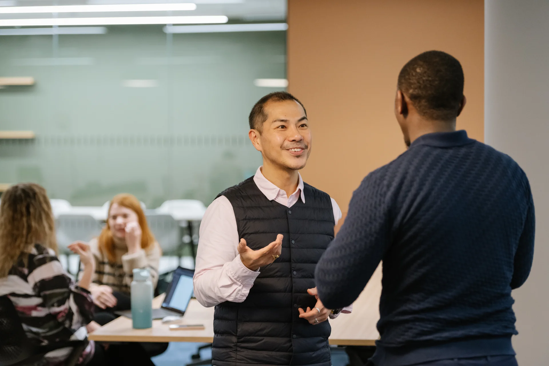 Two men converse in a modern office space, with others seated at desks in the background, under bright indoor lighting.