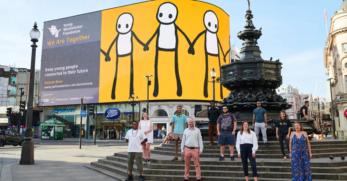 A group of people standing on the steps in a public square in front of a large yellow billboard ad for the **Young Westminster Foundation** campaign, "**We Are Together**."