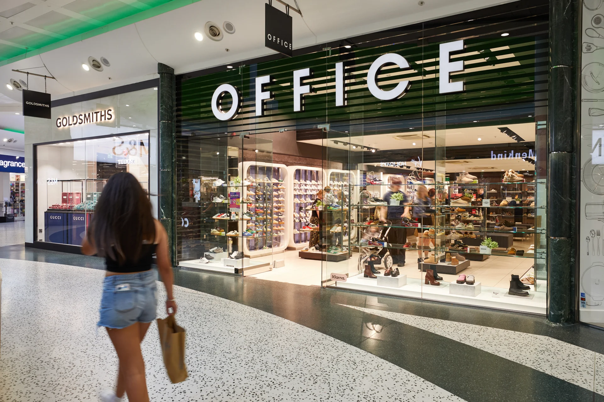 Interior view of the **White Rose Shopping Centre** mall, showing the brightly lit storefronts of the **OFFICE** shoe store and **GOLDSMITHS** jeweler, with a shopper walking past.