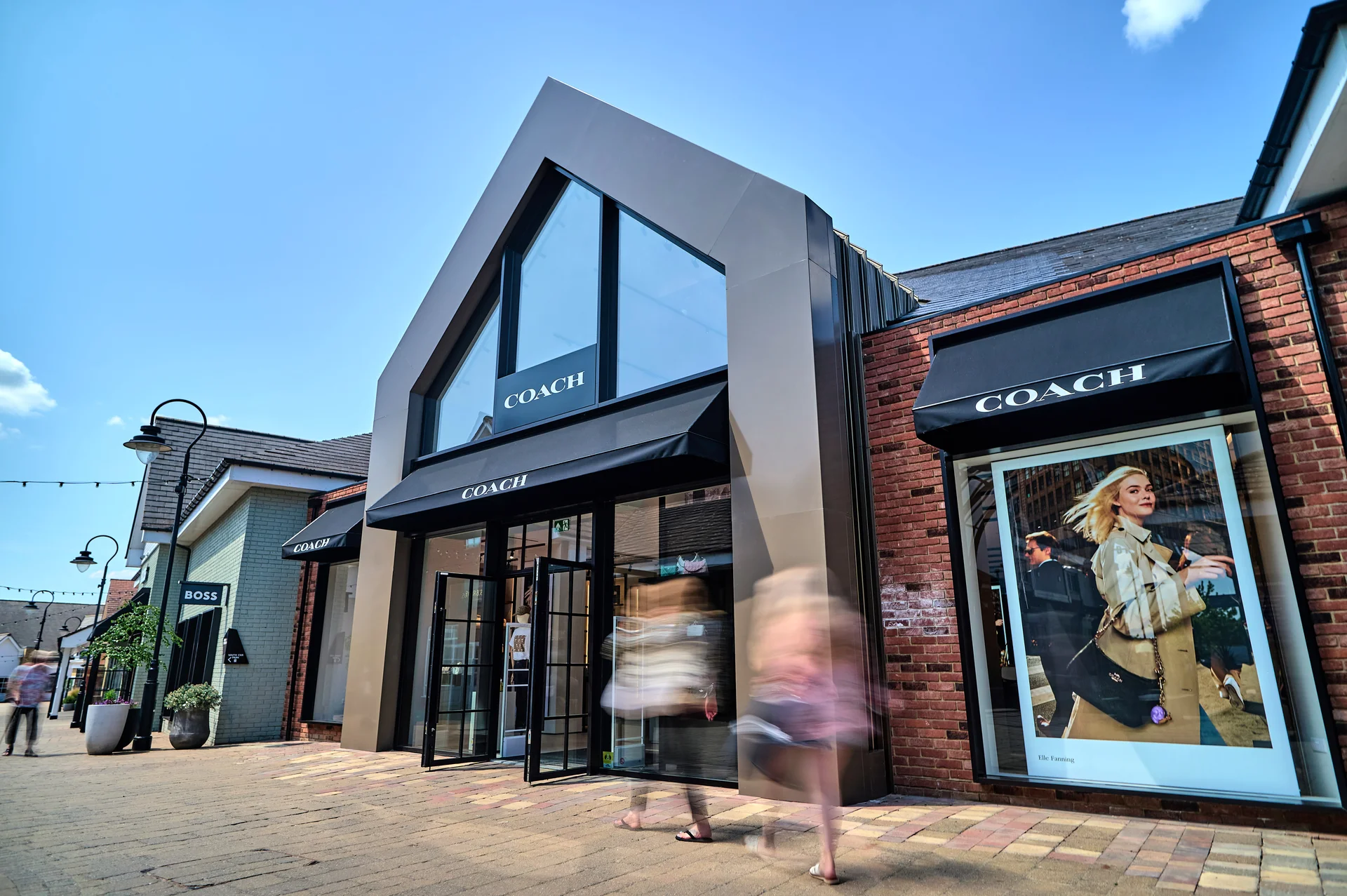 The modern, pitched-roof **Coach** store at **Braintree Village** outlet, featuring dark awnings, large glass windows with advertising, and motion-blurred shoppers walking along the outdoor brick pavement.