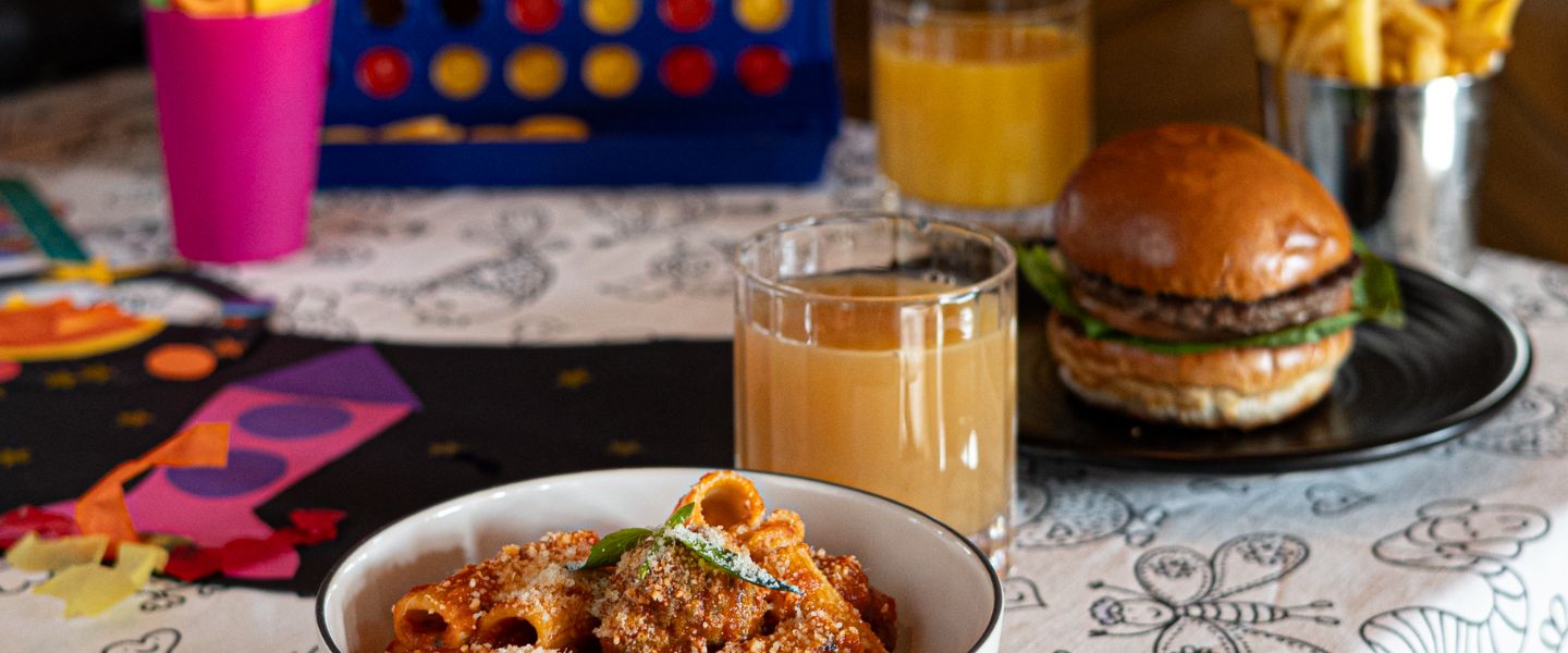 A bowl of pasta on a table next to a burger, all in front of games and Connect 4.