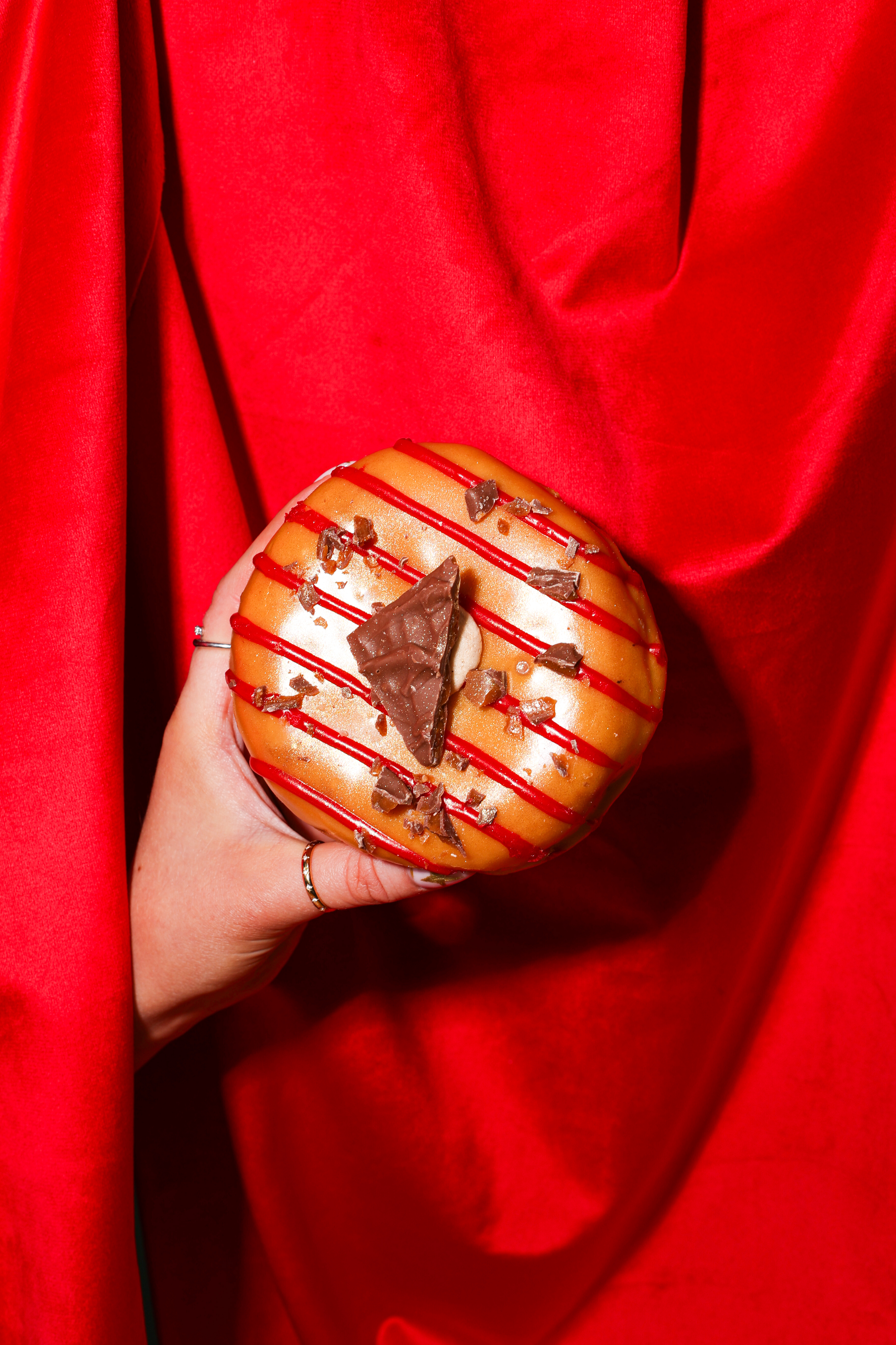 A hand holding a new daim bar doughnut dispalyed against a red background that looks like fabric, the doughnut is glazed with caramel and has sprinklings of a daim bar on top and red stripes 