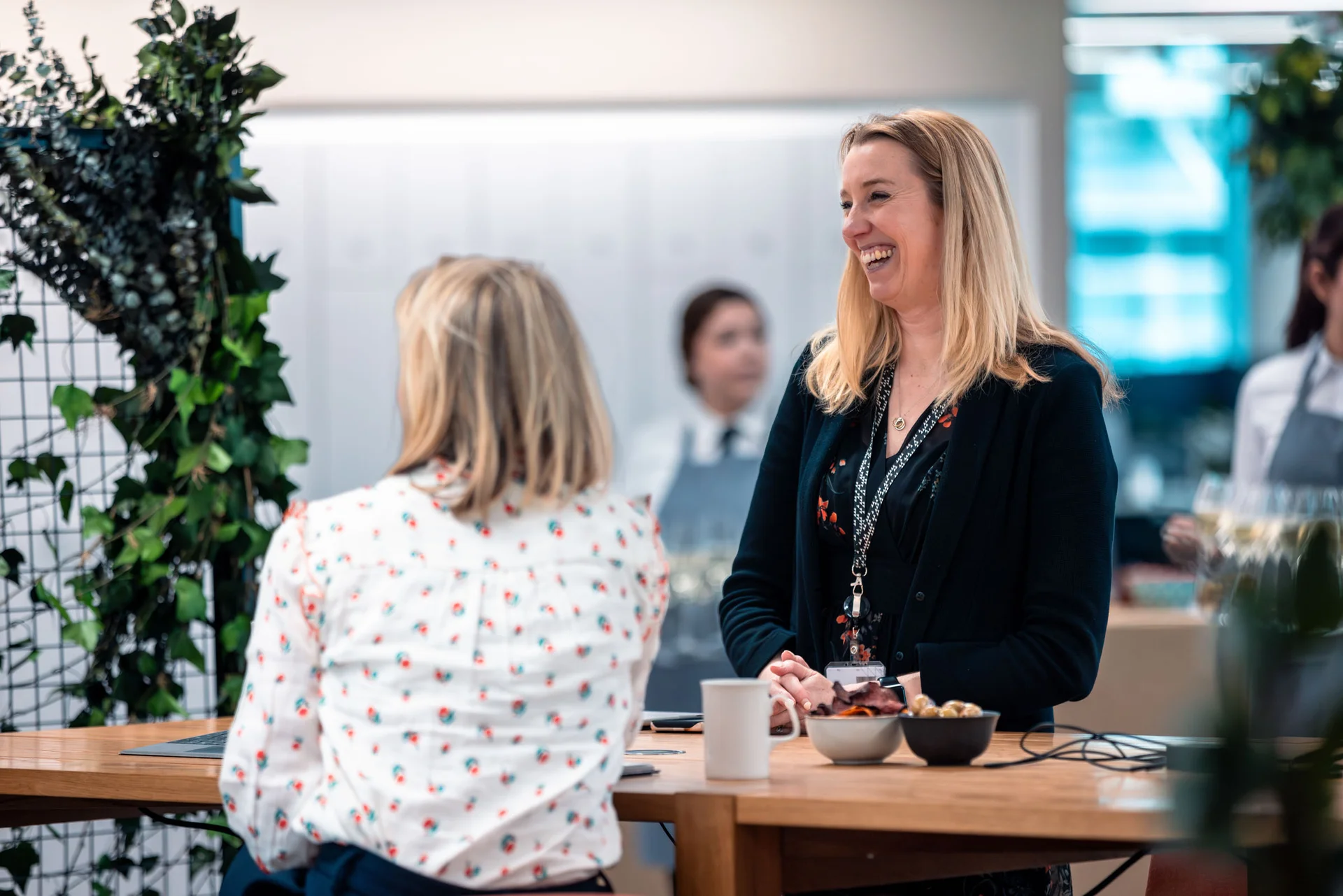Two women converse at a wooden table with snacks and drinks, in a modern, well-lit space with greenery and blurred background figures.