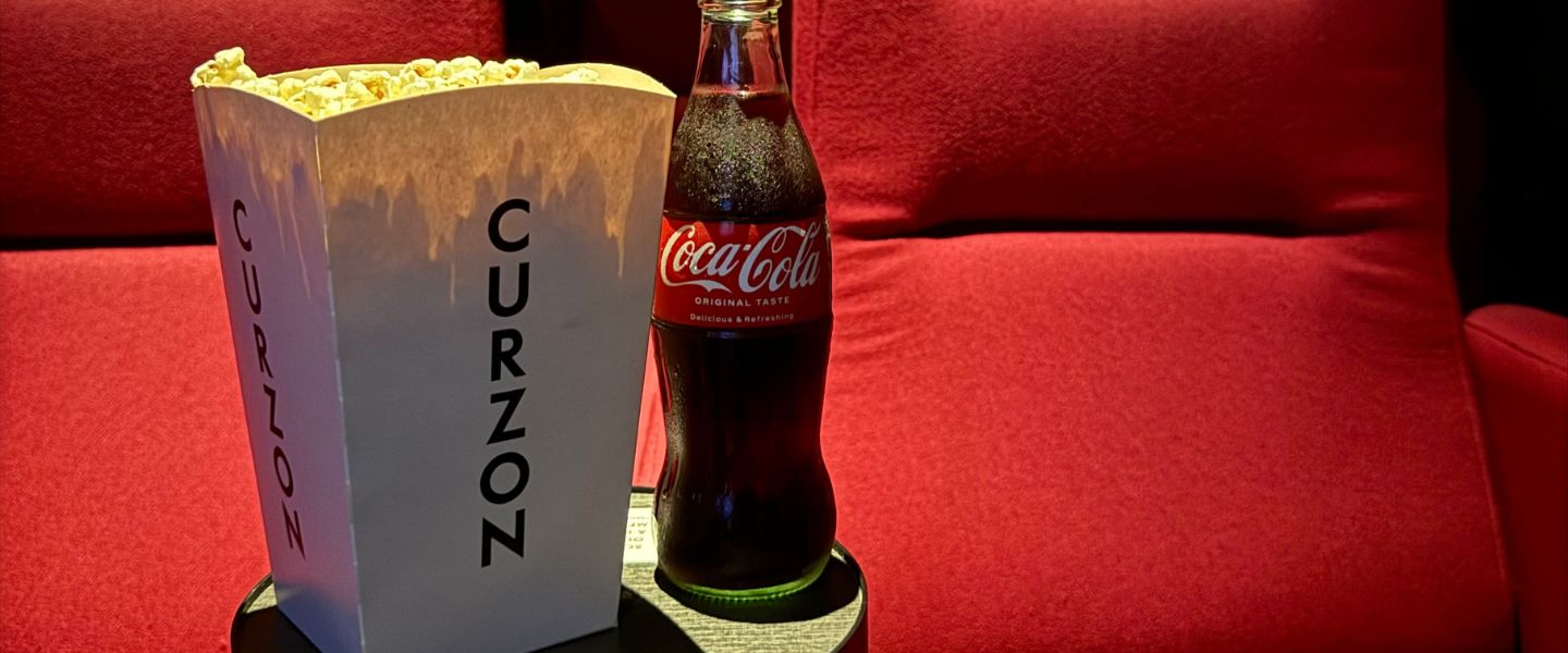 A carton of popcorn from Curzon next to a glass bottle of Cola against red cinema seats