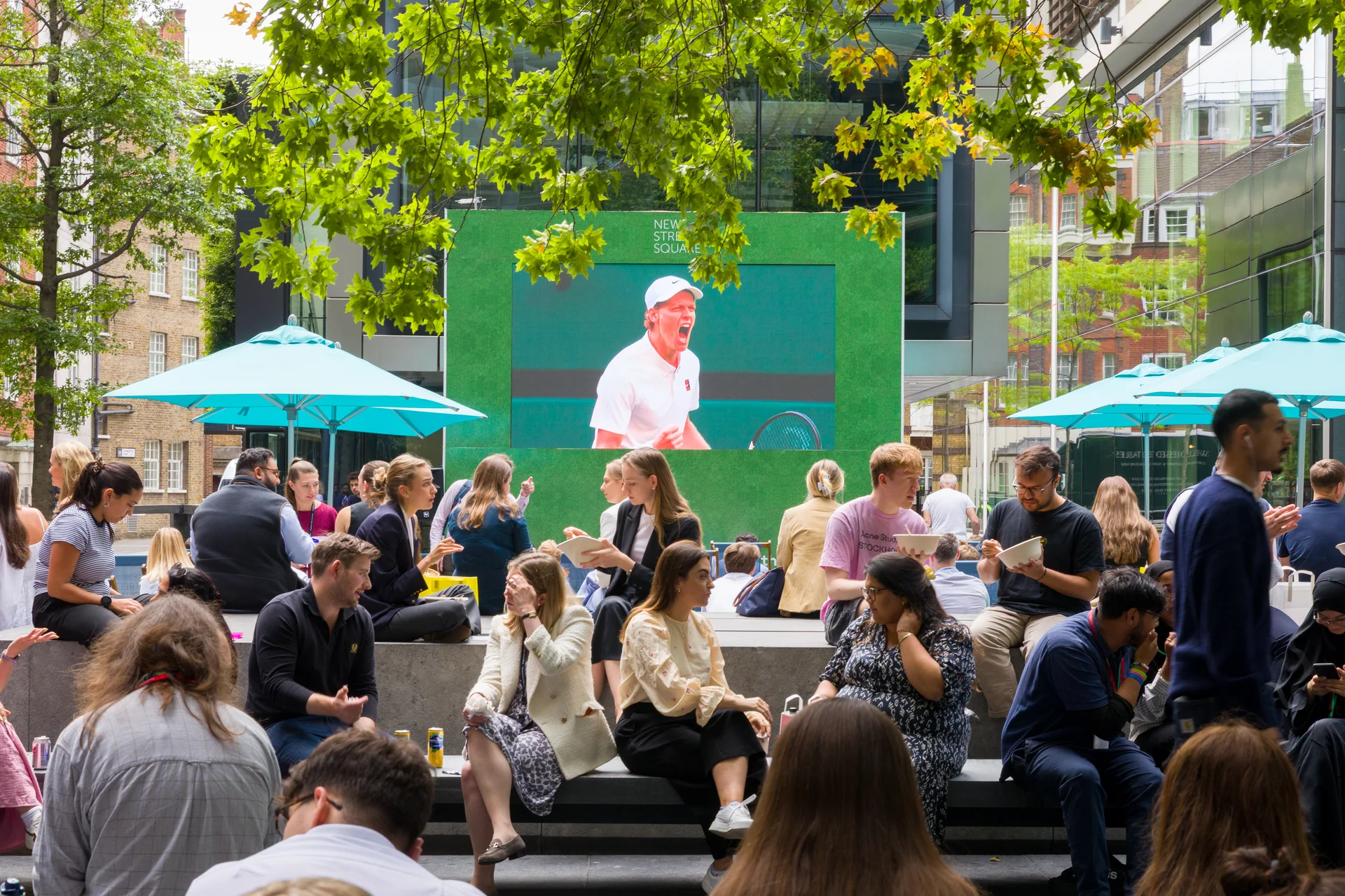 Outdoor public event with people watching tennis on a large screen under trees and blue umbrellas