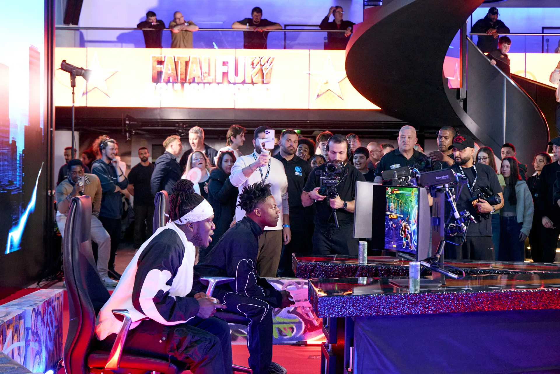 Gaming event with players competing on stage, surrounded by spectators and press.