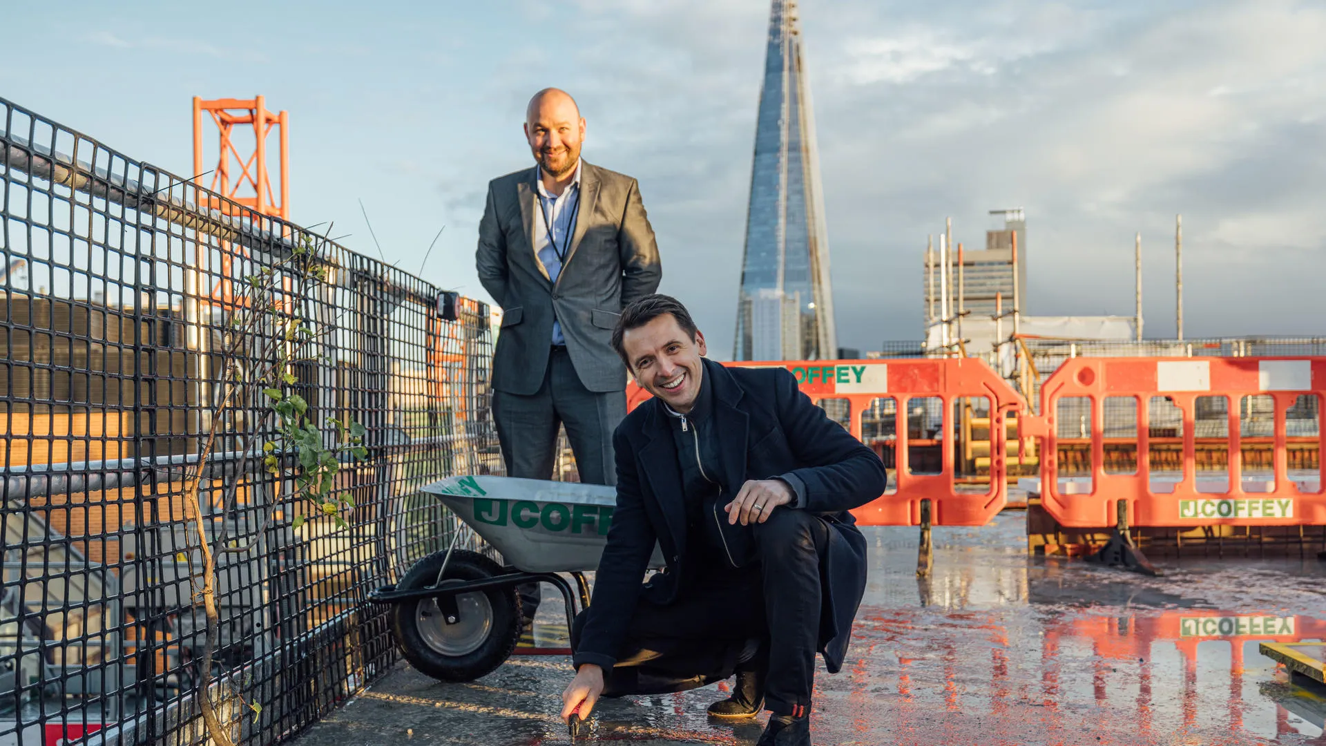 Two men at a construction site with the Shard in the background, one kneeling and one standing behind a wheelbarrow, with "J.COFFEY" visible.