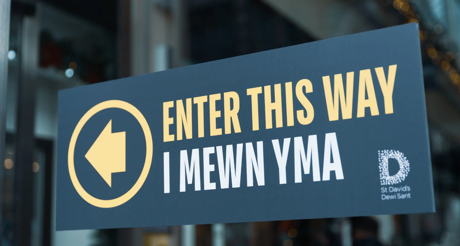 Blue sign with "ENTER THIS WAY" and "St David's" text, and a left arrow, on a glass door.