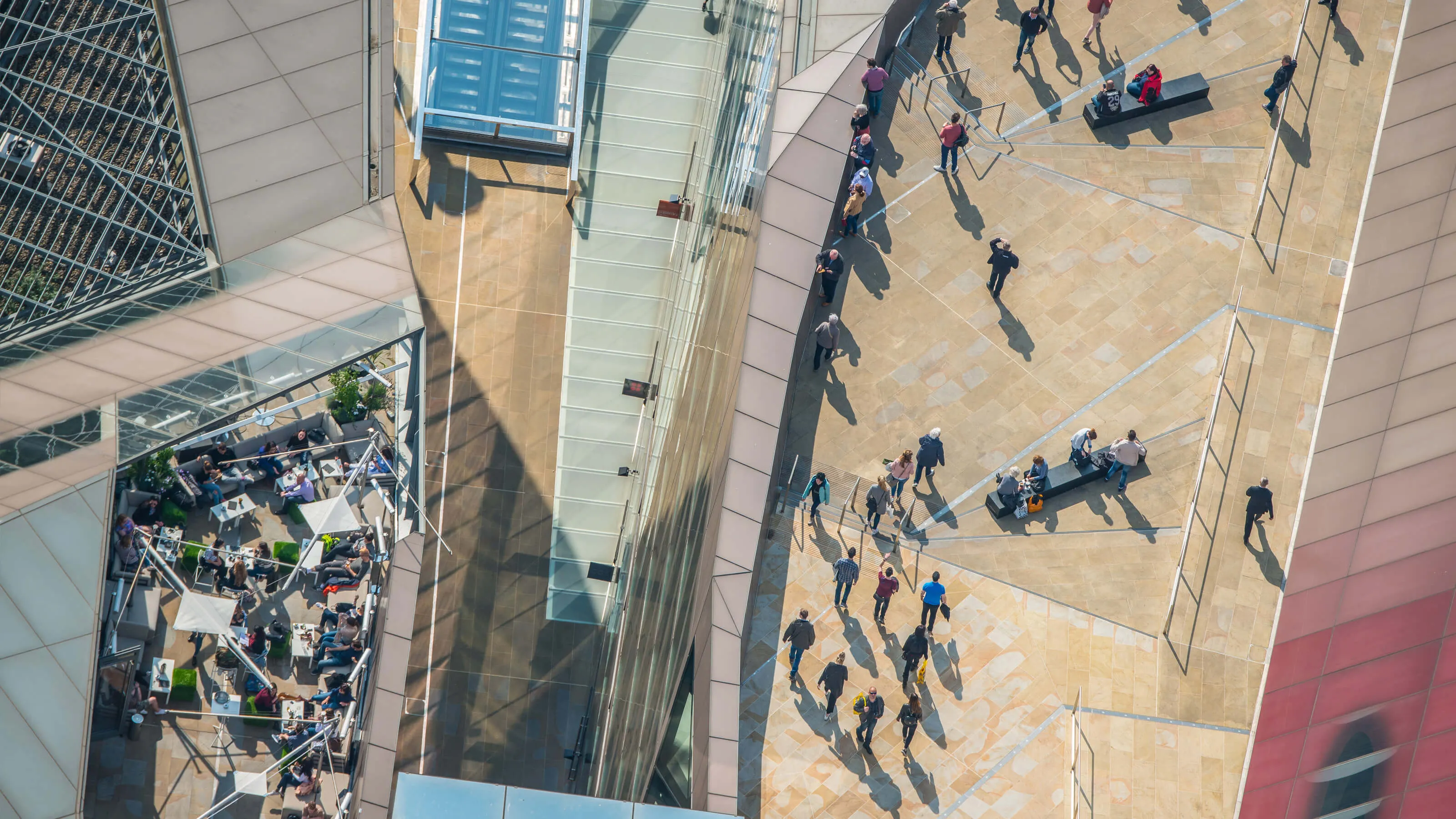 A top-down view of a sunny city plaza or terrace, with people walking and casting long shadows.