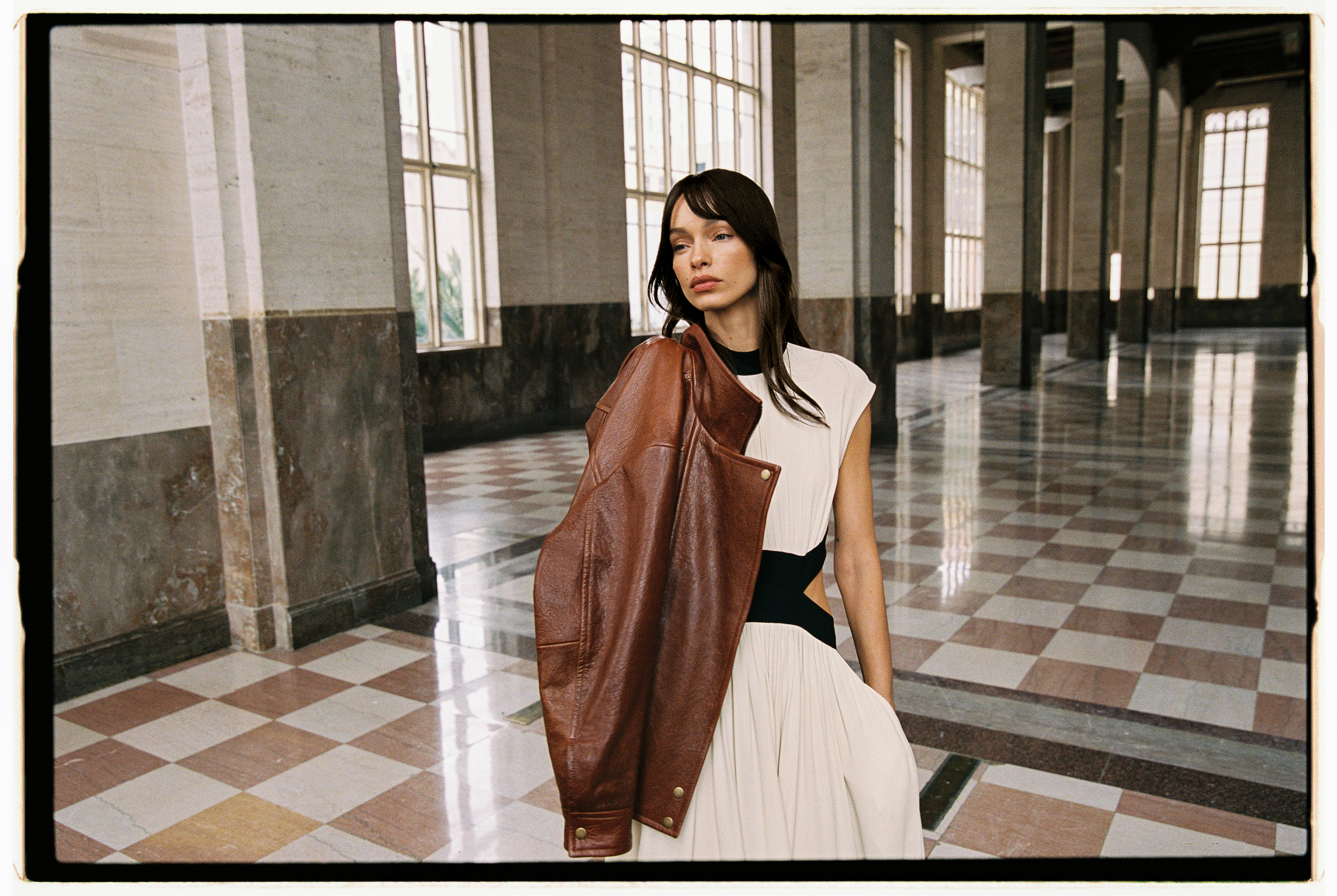 A woman in a white dress with a leather jacket slung over one shoulder stands in a large hall space with a brown and white chequered tile floor and large windows.