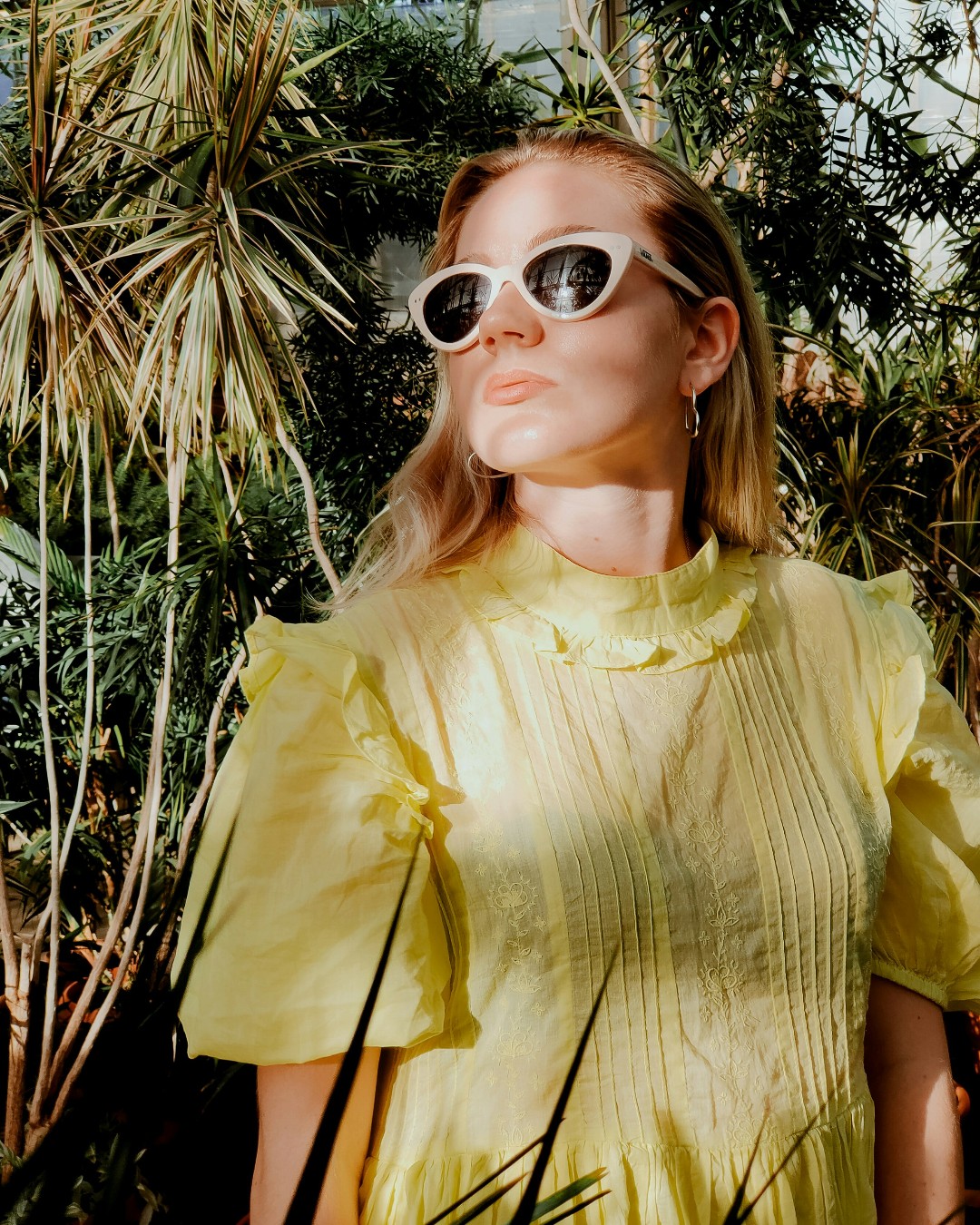 A blonde woman dressed in a yellow top and sunglasses stands among tropical plants.